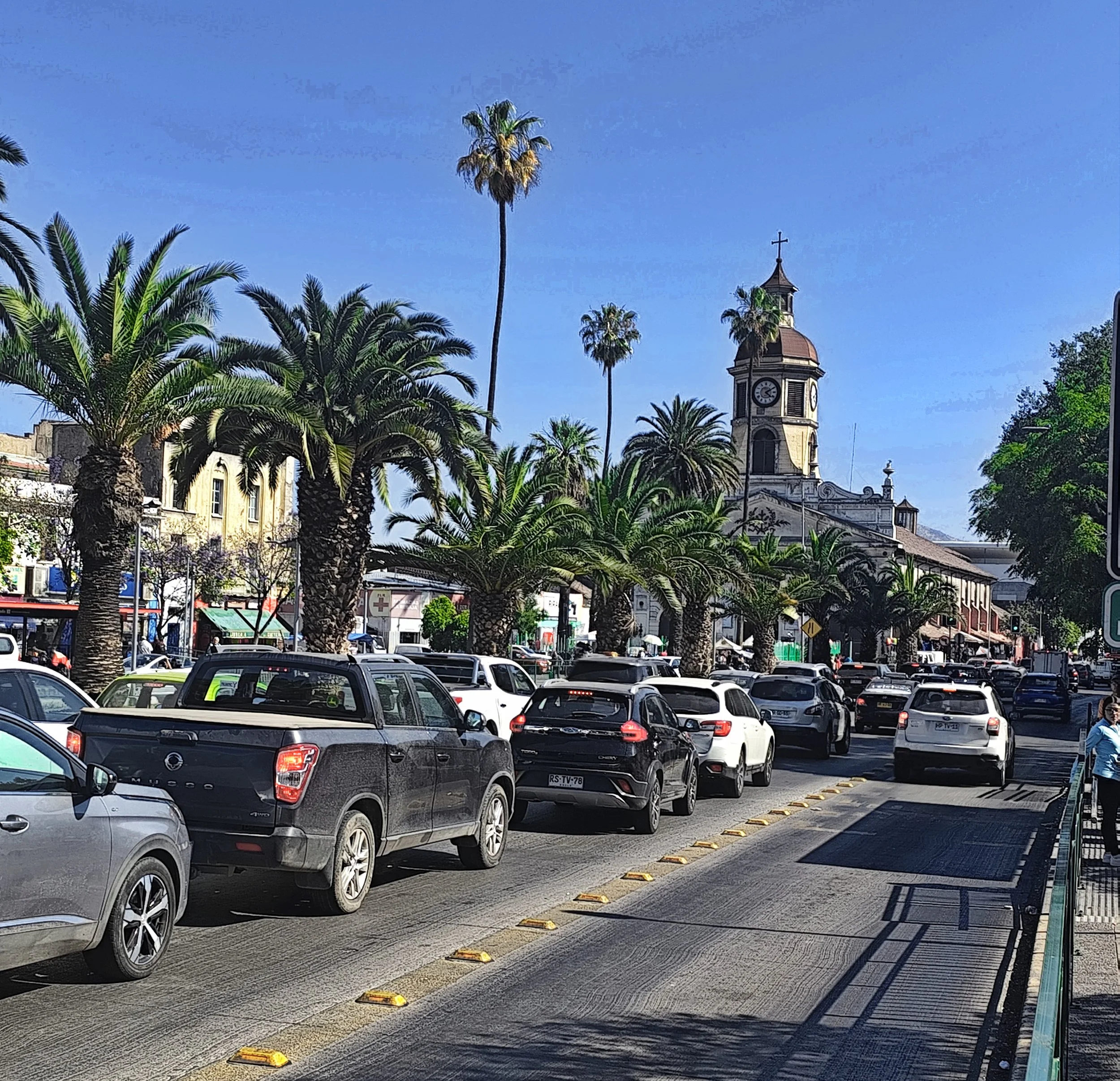 A busy street scene with palm trees, parked and moving cars, a church with a clock tower, and pedestrians under a clear blue sky.