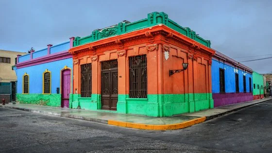 Colorful corner building with orange, pink, purple, green, blue, and teal sections, featuring barred windows and decorative architectural details, on a city street.