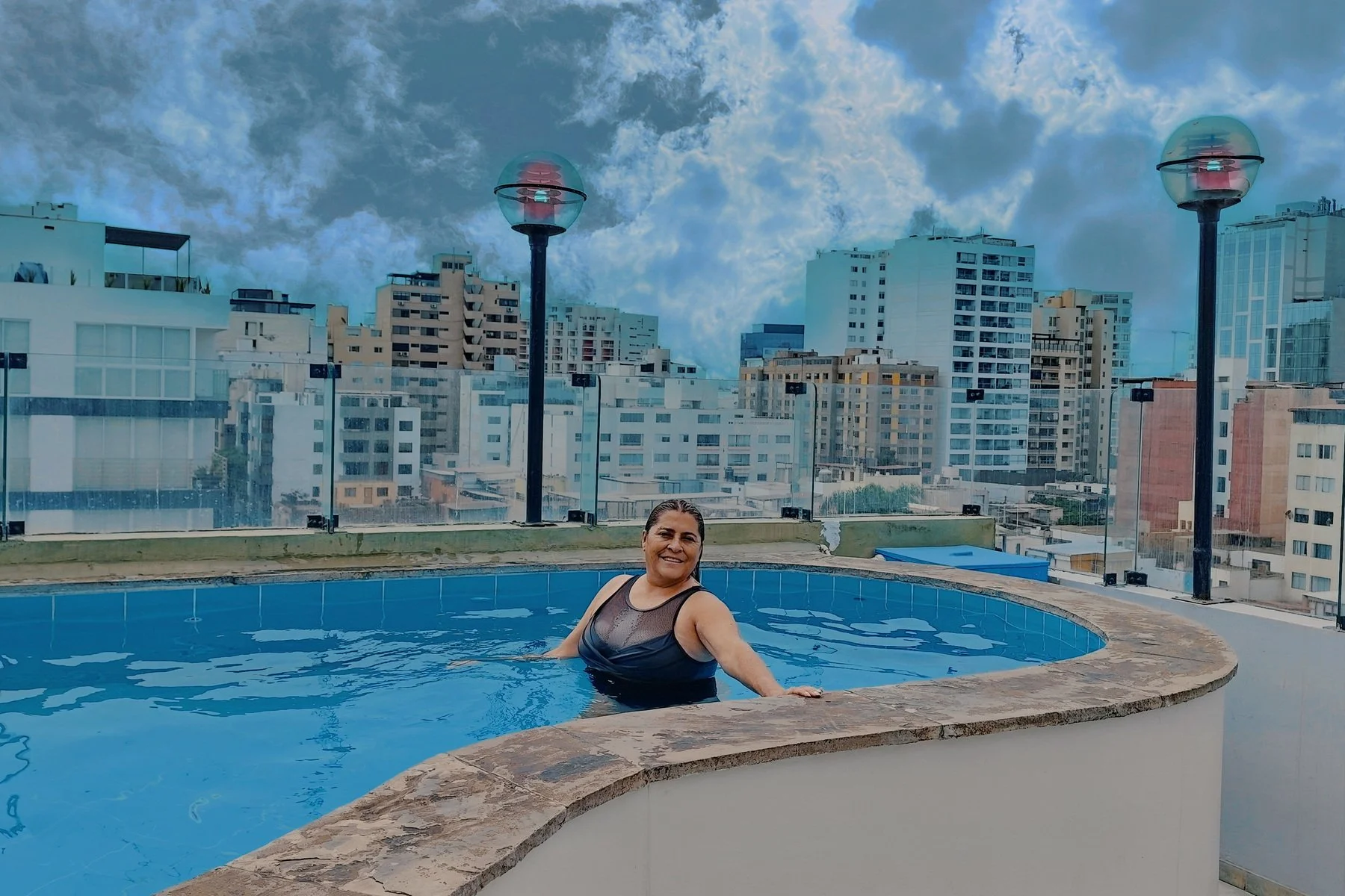A woman in a black swimsuit smiling while standing in a rooftop pool with a city skyline and cloudy sky in the background.