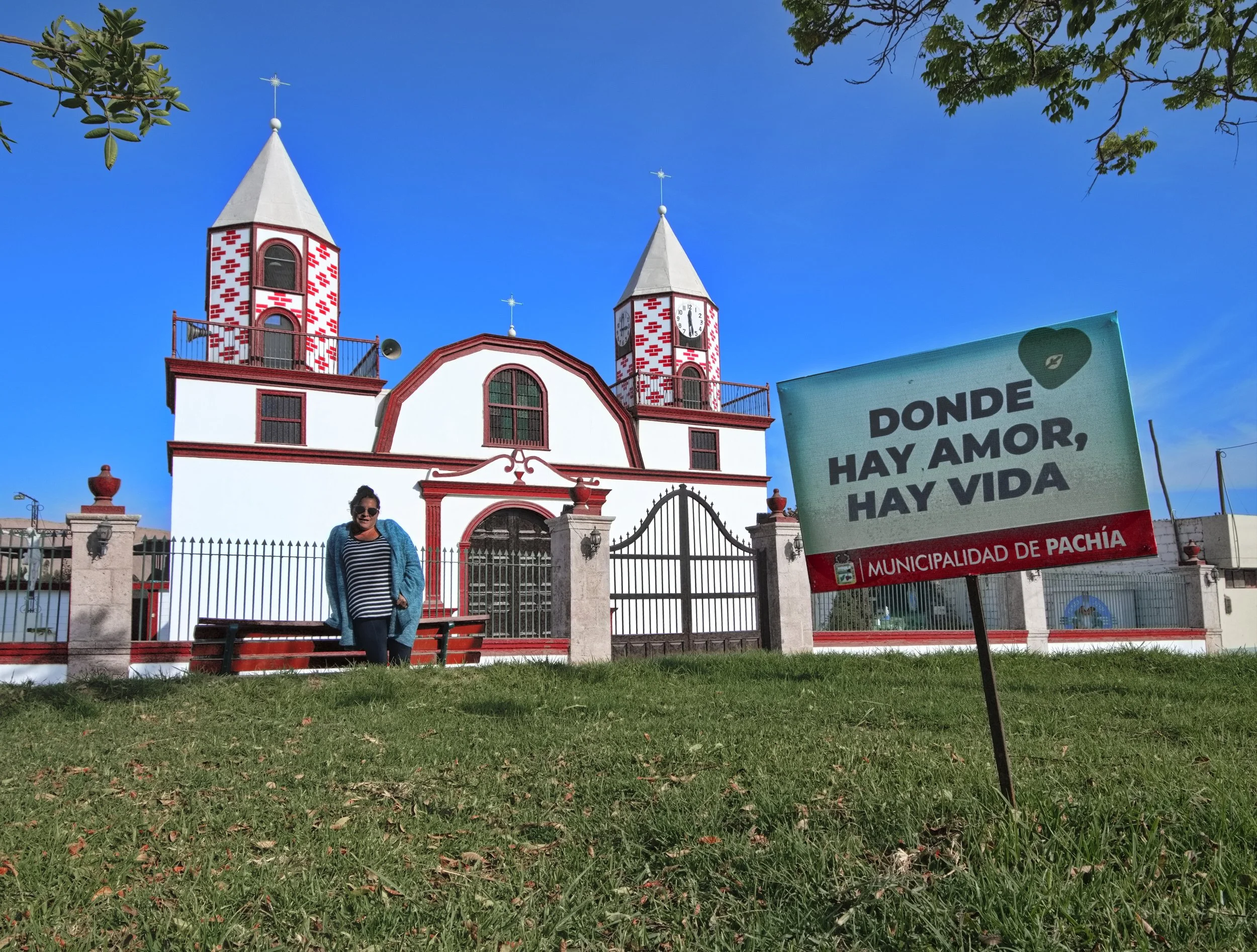 A woman standing on a bench in front of a white church with red accents and twin clock towers with blue skies overhead. A sign in the foreground reads "Donde hay amor, hay vida" from the Municipalidad de Pachía.