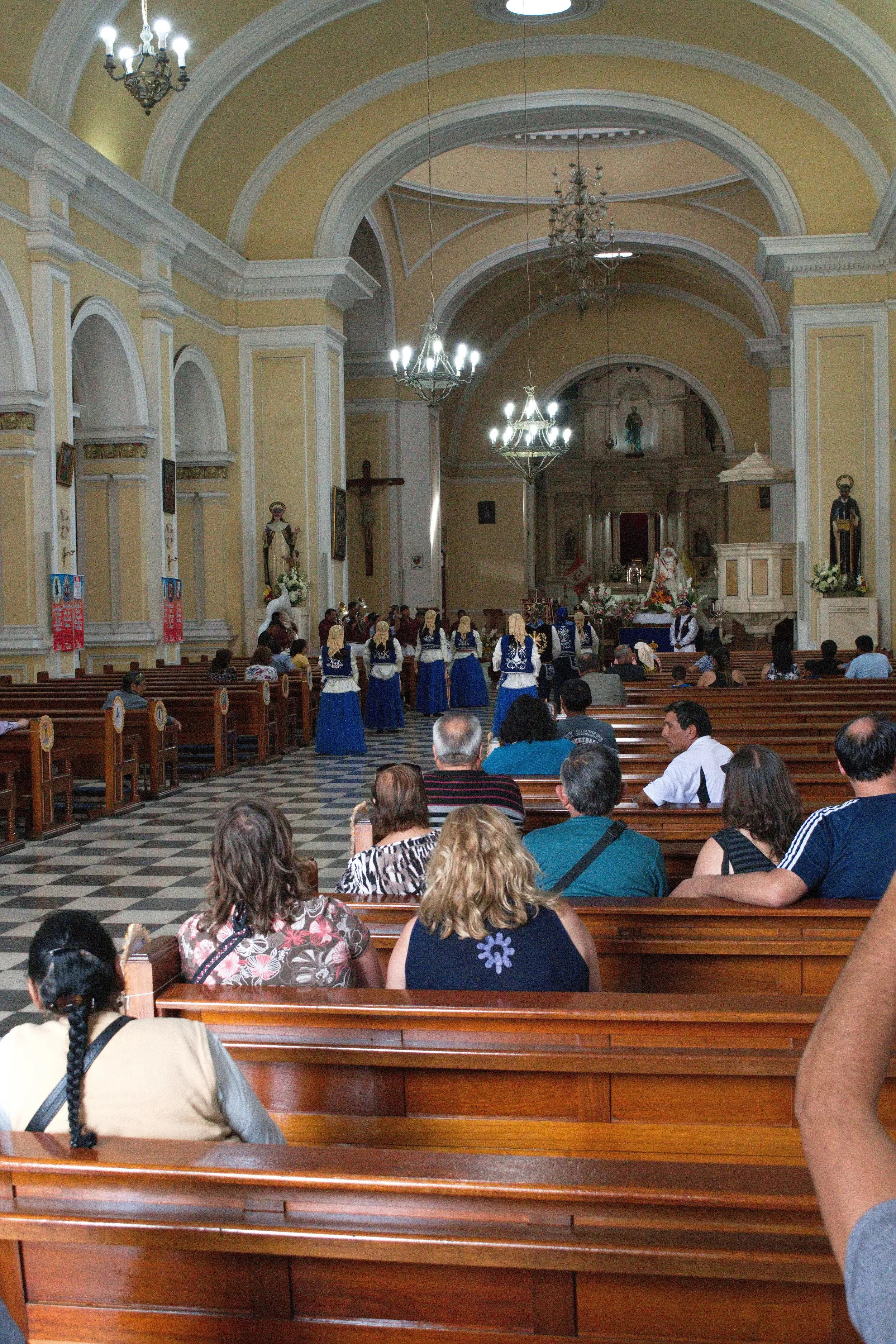 People seated inside a church, watching a cultural dance performed by women in traditional blue and white dresses, with an altar decorated with flowers and religious statues at the front.