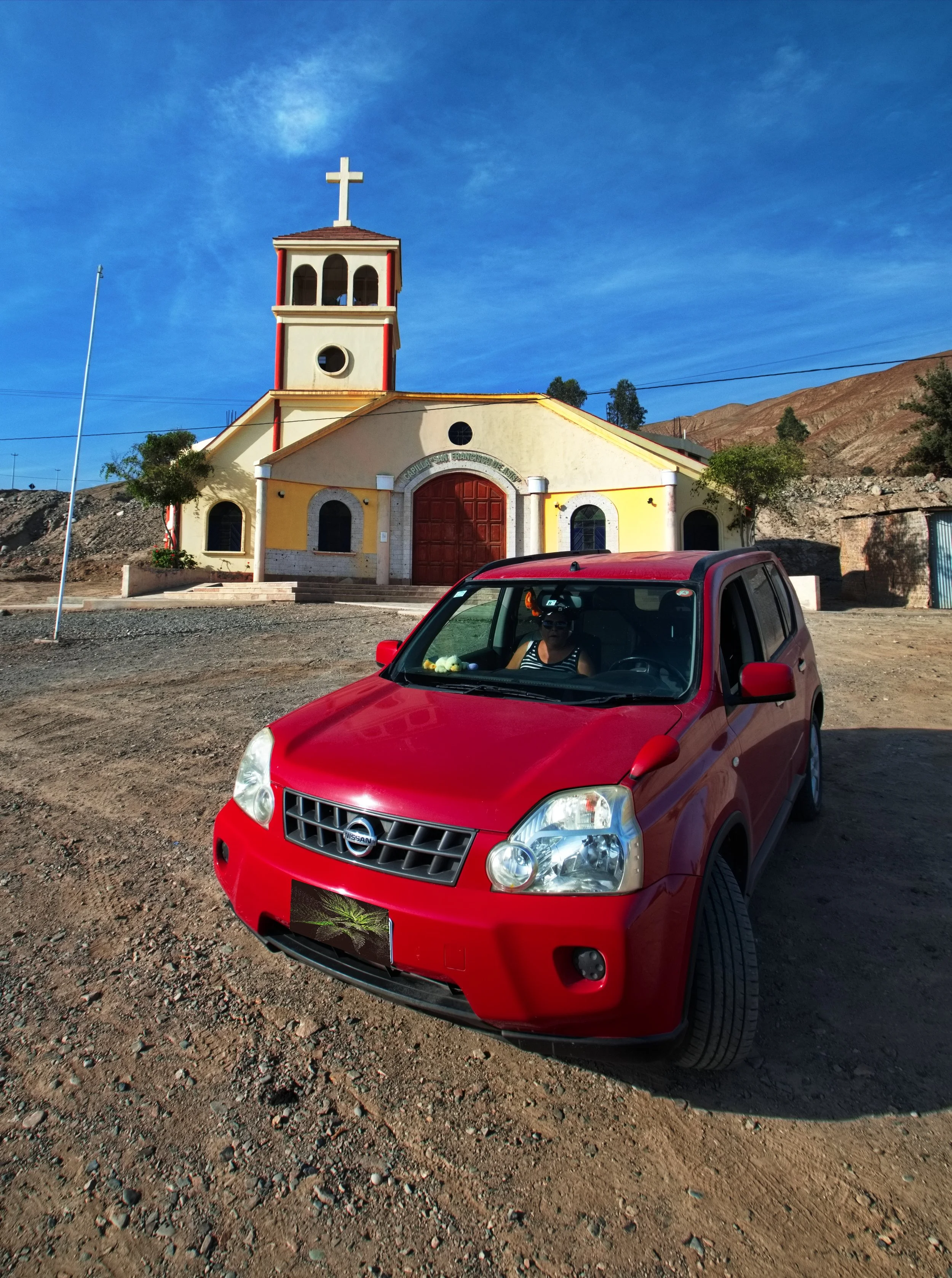 A red Nissan SUV parked on a dirt area in front of a small church with a cross on top, blue sky overhead, and mountains in the background.
