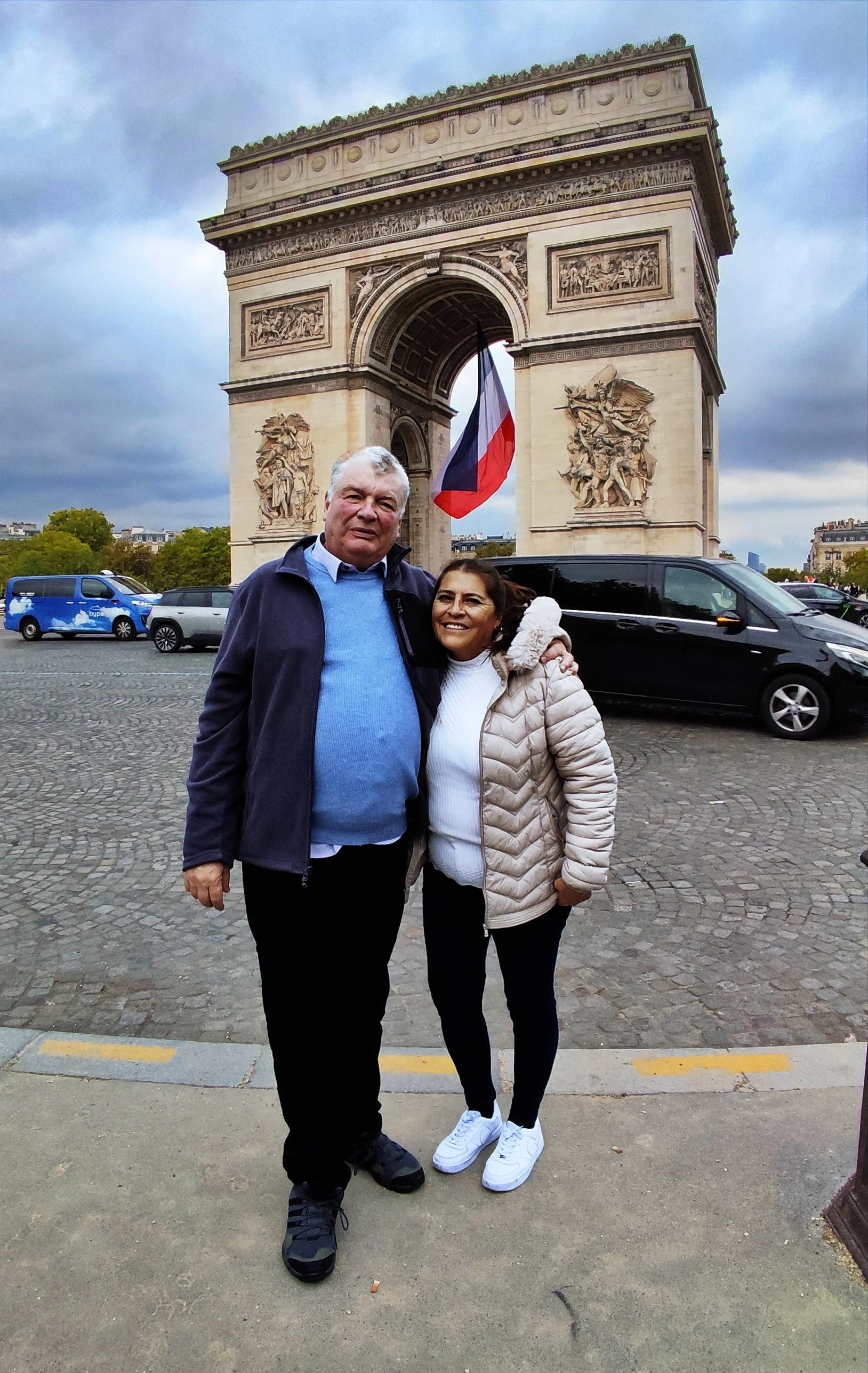 Two people standing in front of the Arc de Triomphe in Paris, France, with a French flag hanging from the arch, during cloudy weather.