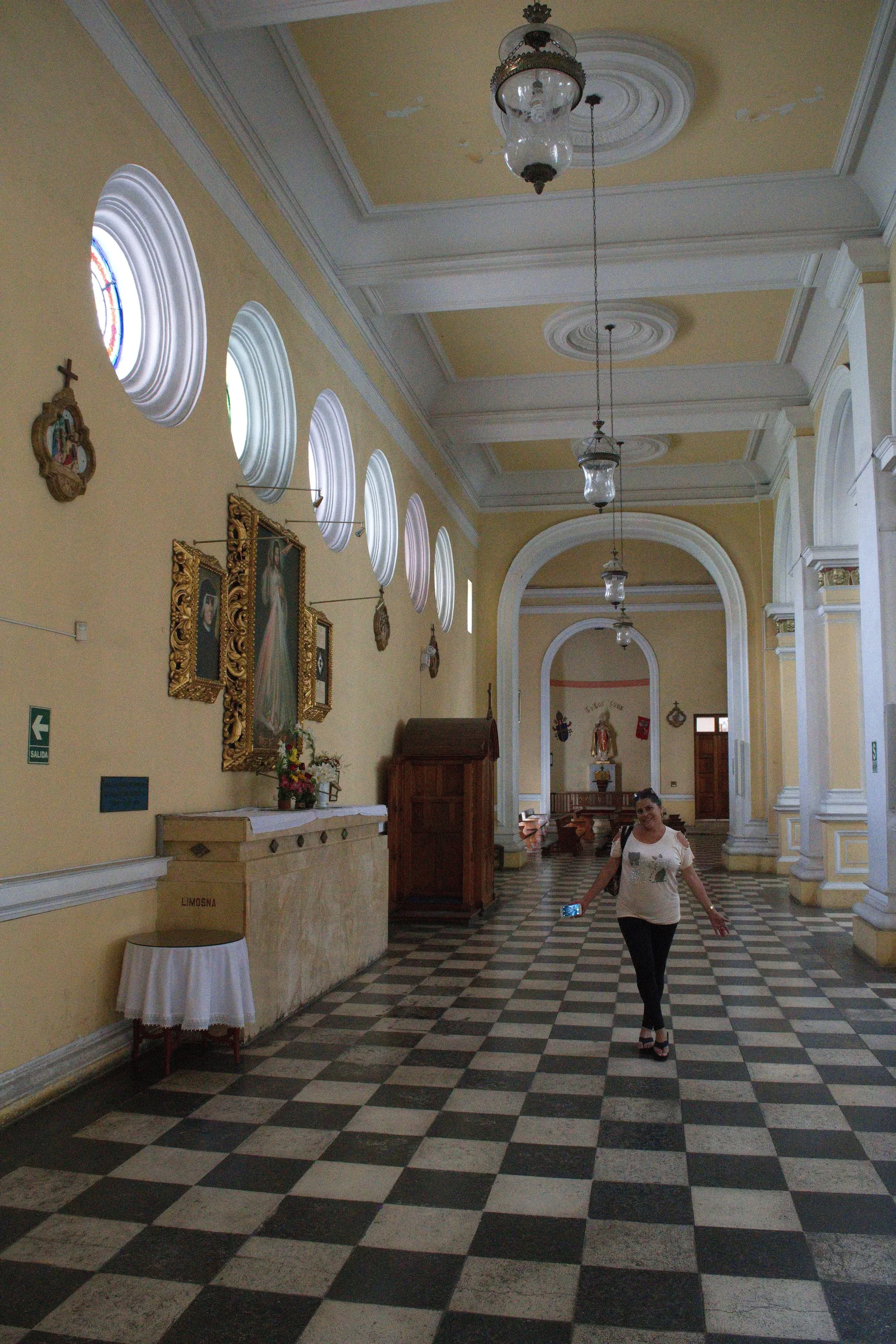 Interior of a church with checkered tile floors, yellow walls, stained glass windows, religious icons, and a woman walking down the aisle.