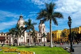 A historic church with domed towers in a park, featuring palm trees, a lamppost, and colorful buildings in the background under a blue sky.