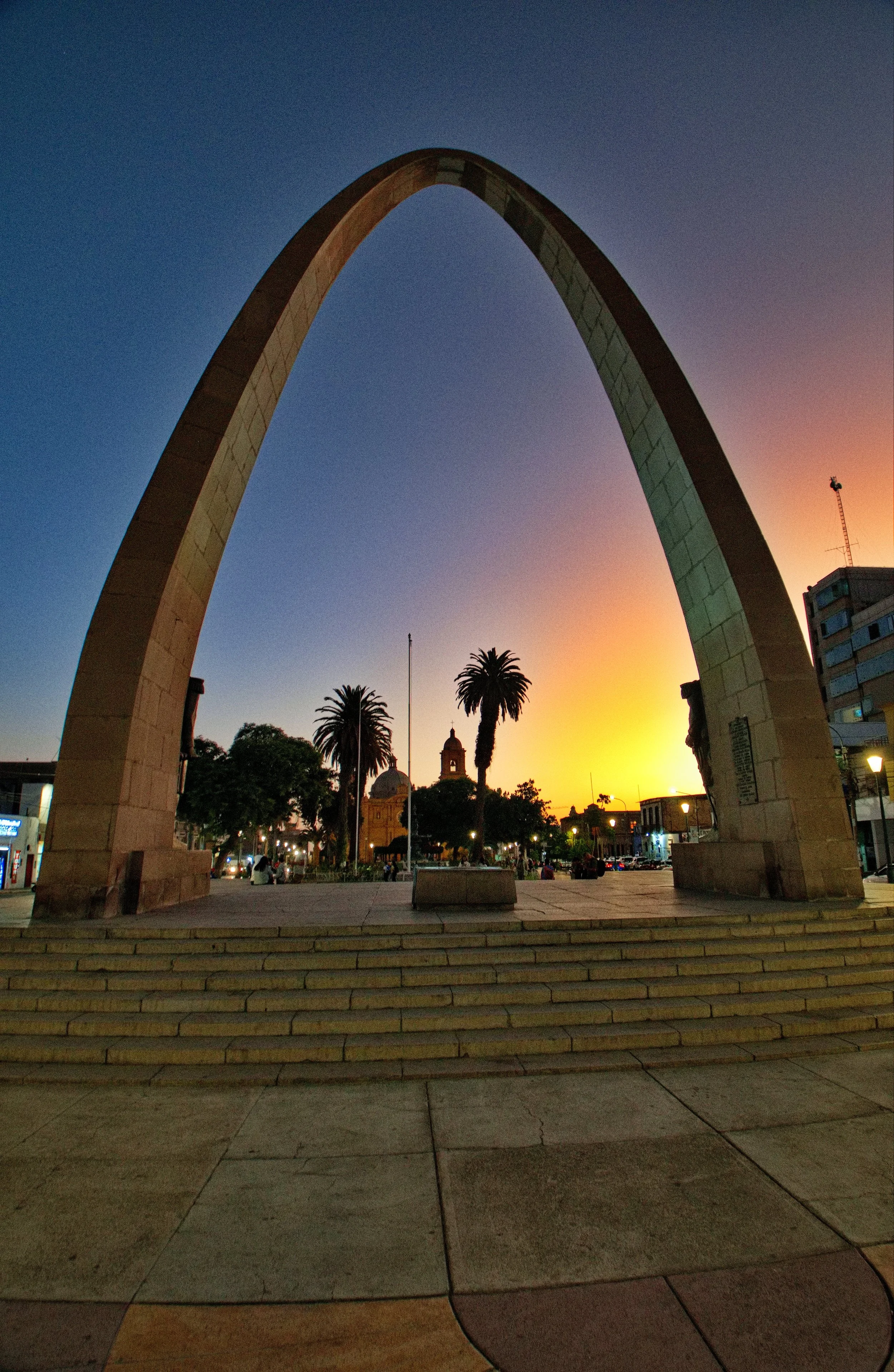 The Gateway Arch at sunset in St. Louis, Missouri, with steps leading up to it, palm trees in the background, and a colorful sky.