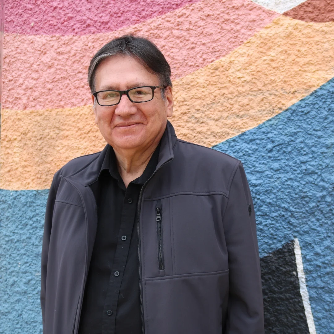 A man with glasses and a black jacket smiling in front of a colorful, textured mural with pink, orange, and blue sections.