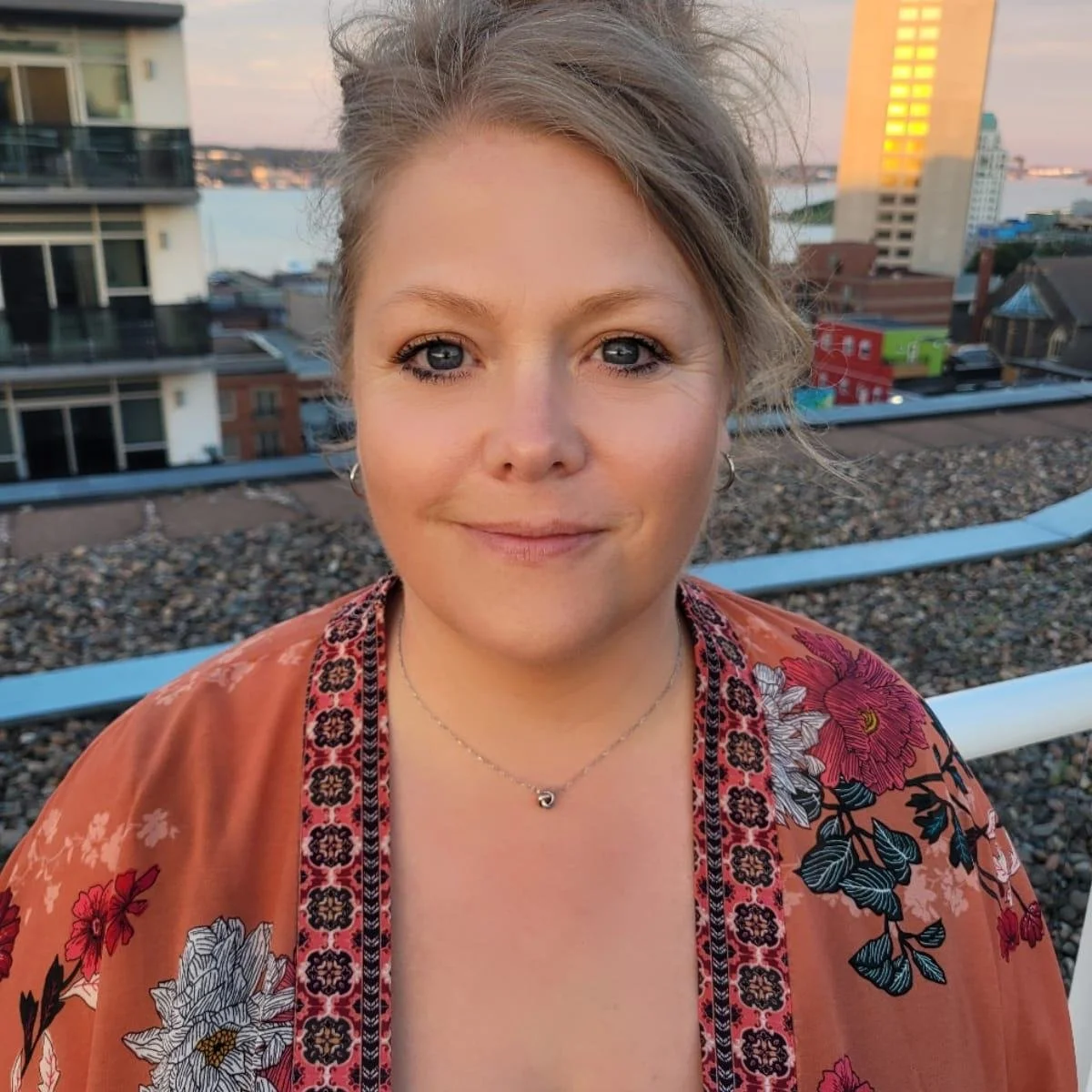 A woman with light brown hair and blue eyes taking a selfie on a rooftop during sunset, wearing a floral patterned top and a necklace with a small pendant, with city buildings and water in the background.