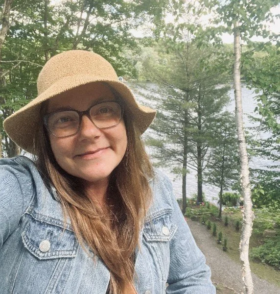 A woman in a denim jacket and a straw hat smiling, standing outdoors near a lake with trees, on a cloudy day.