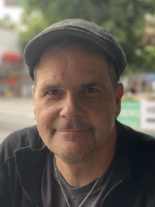 A man with a gray cap and a black shirt smiling outdoors on a sunny day.
