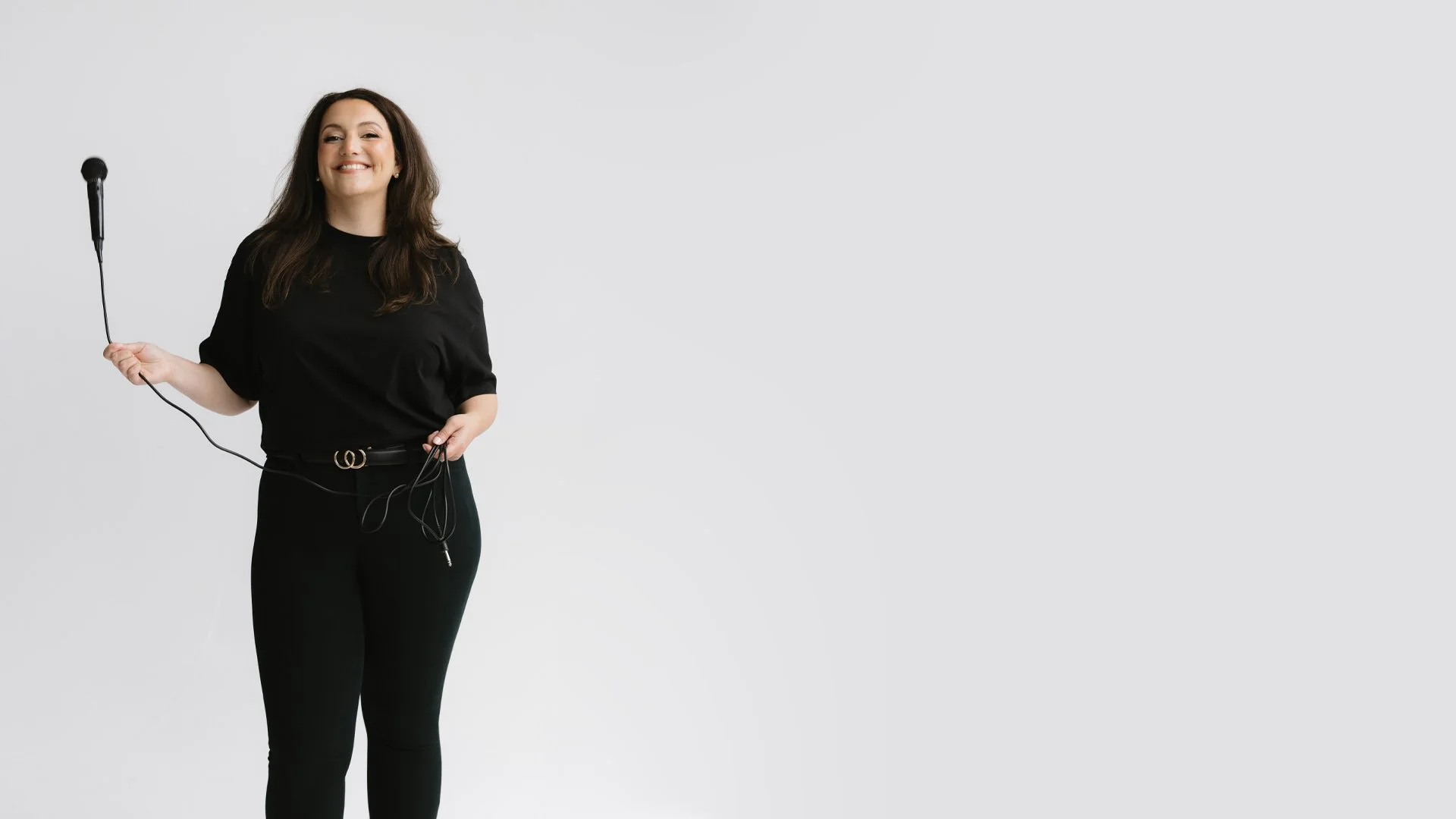 Jessica Flynn  in black clothes holding a microphone and headphone cable, standing against a plain white background.