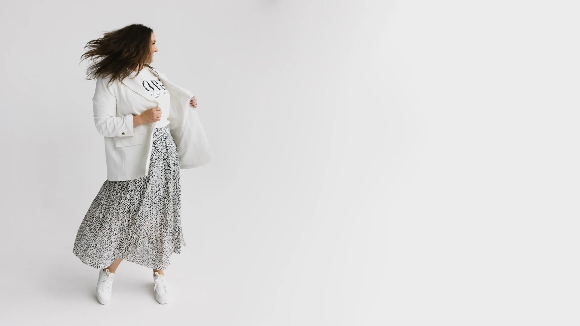 Jessica Flynn, wearing a white blazer, a white T-shirt, a long patterned skirt, and white sneakers, stands against a plain white background, holding open her blazer with one hand.