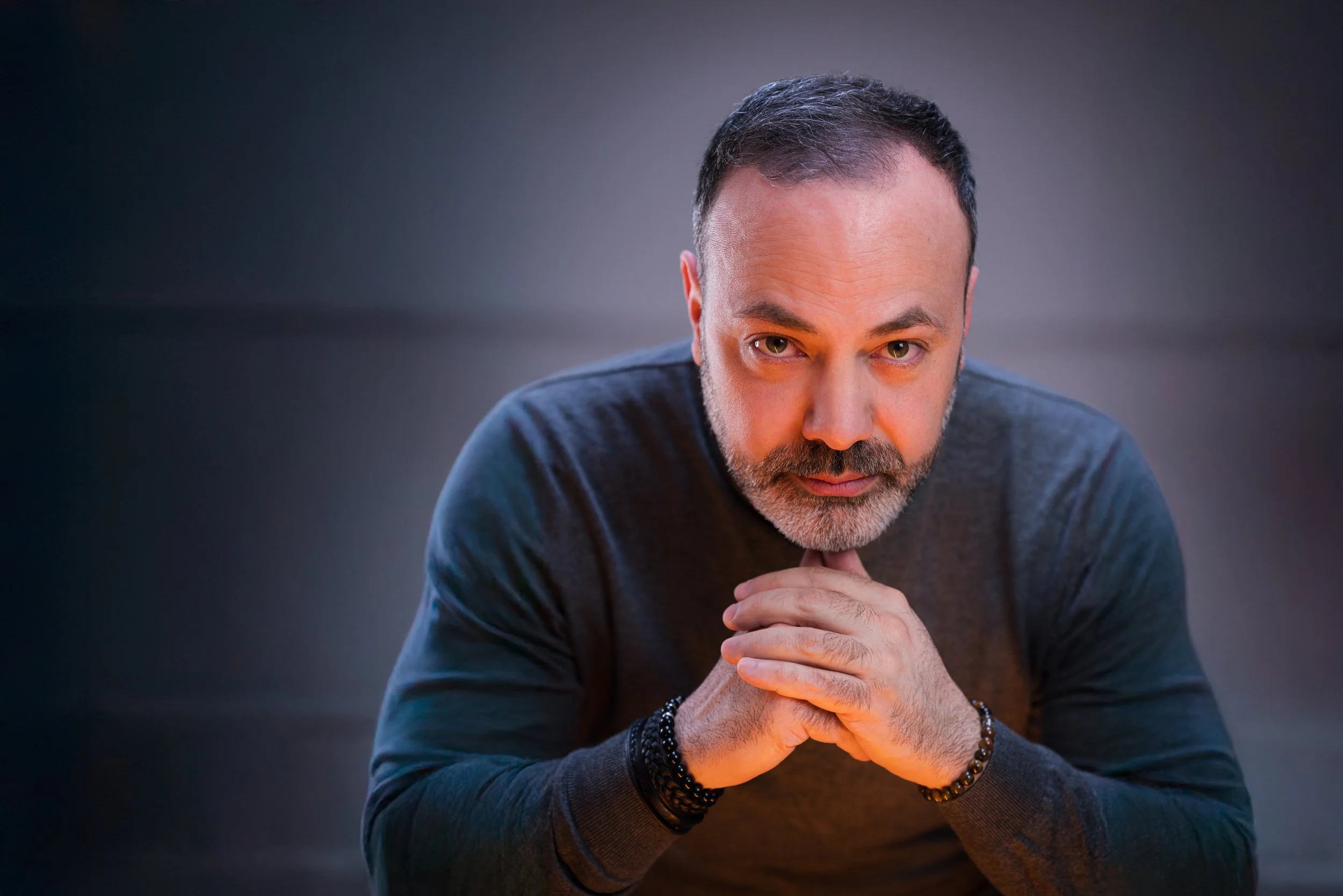Nahaufnahme eines Mannes mit grauem Bart in dunklem Pullover, der nach vorne blickt und die Hände vor seinem Gesicht hat, vor dunkelgrauem Hintergrund.