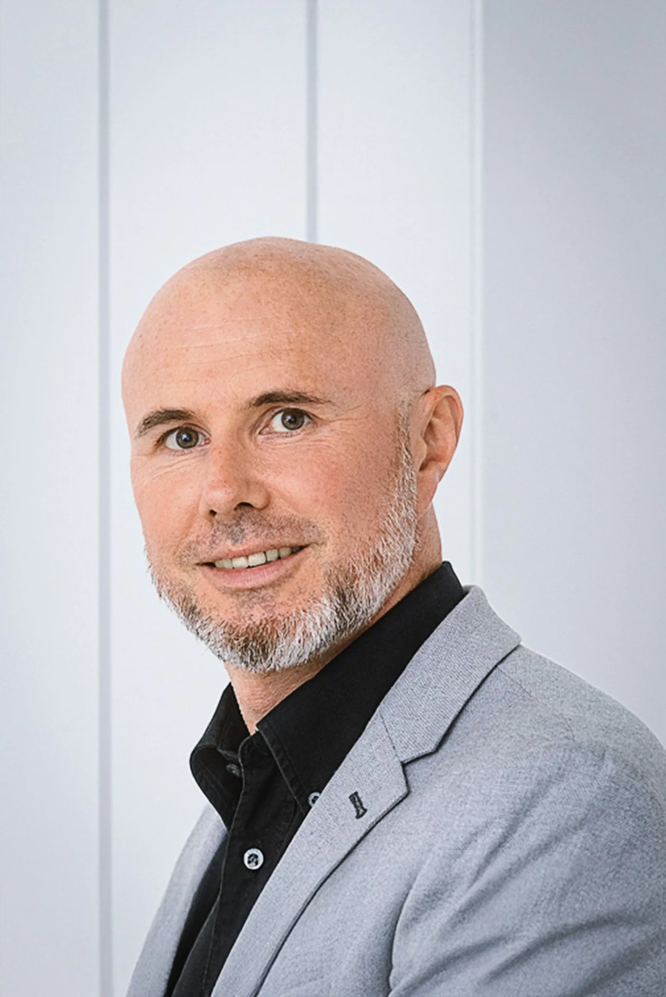 Ein Mann mit Glatze, grauem Bart und grauem Anzug posiert vor einem weißen Hintergrund.