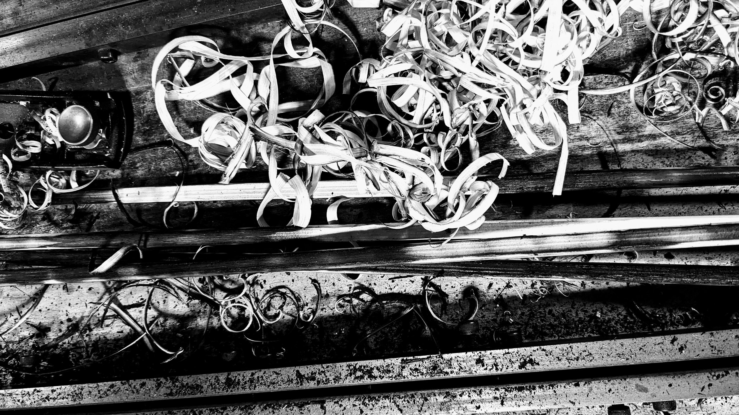 A black and white photo of a cluttered workbench with wood shavings, screws, and tools scattered around, including a measuring tape and a power drill.