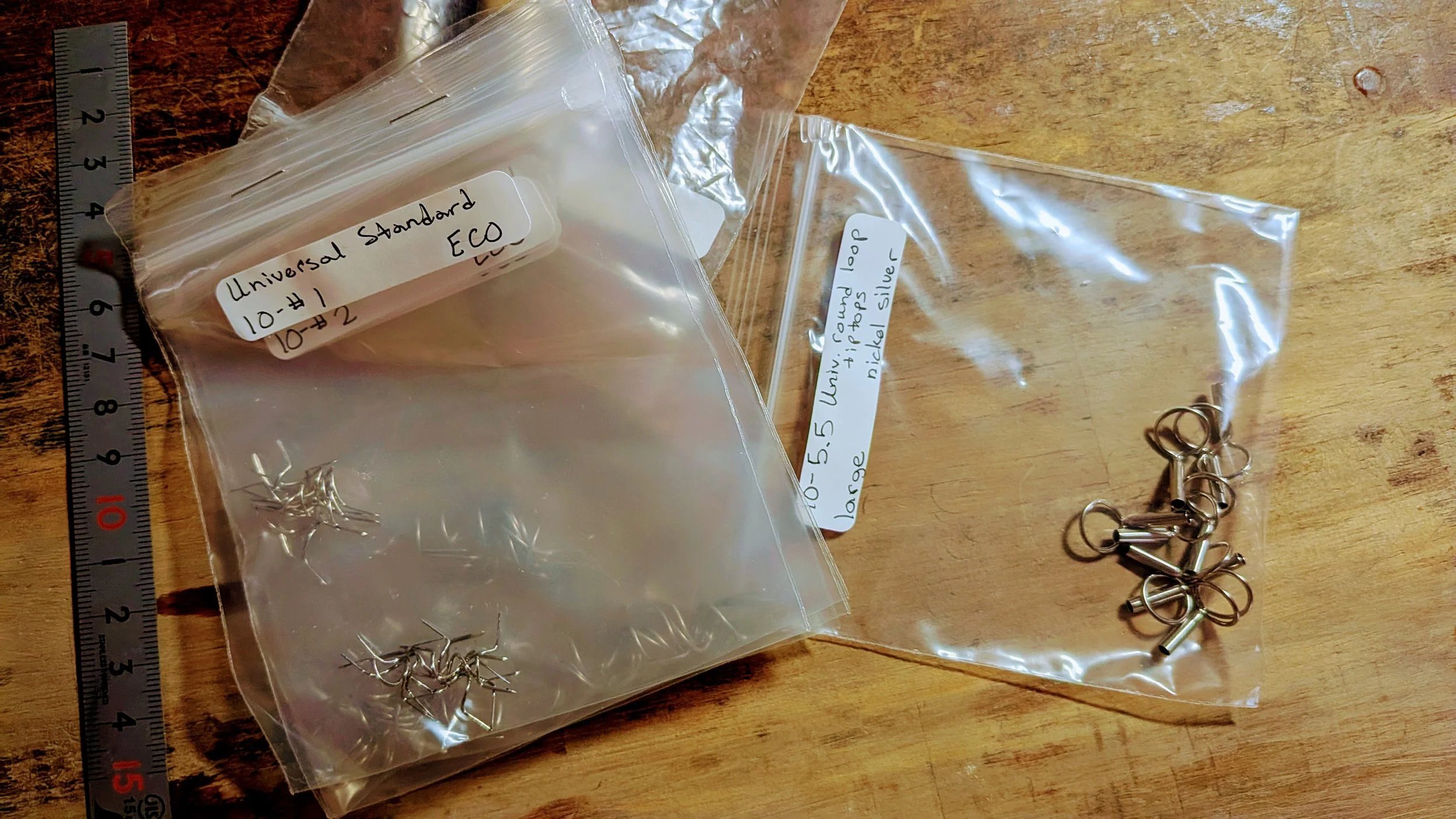 Small plastic bags containing silver jewelry hooks, labeled 'Universal Standard ECO' and '5.5 large'. A metal ruler is on the left side, providing measurement reference.