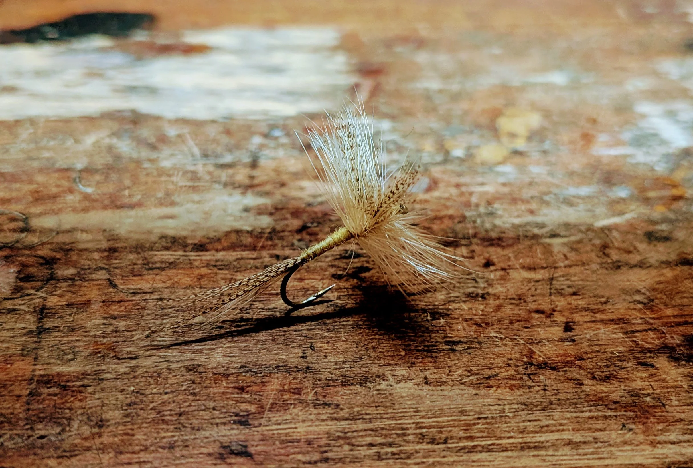 A close-up of a fishing hook with a gray and white feathered lure attached, resting on a wooden surface.