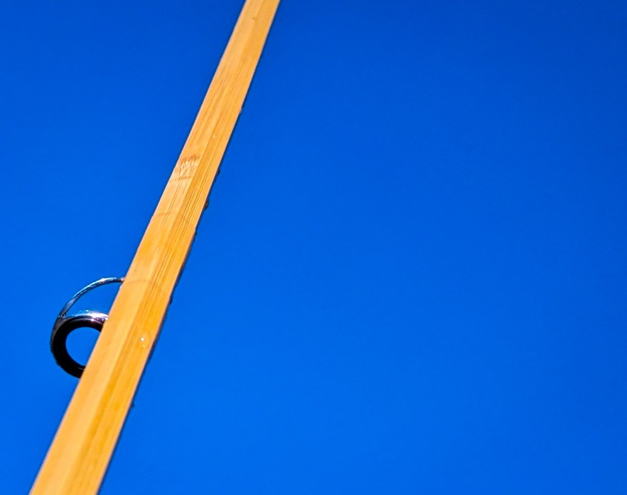 Close-up of a wooden fishing rod with a black reel against a bright blue sky.