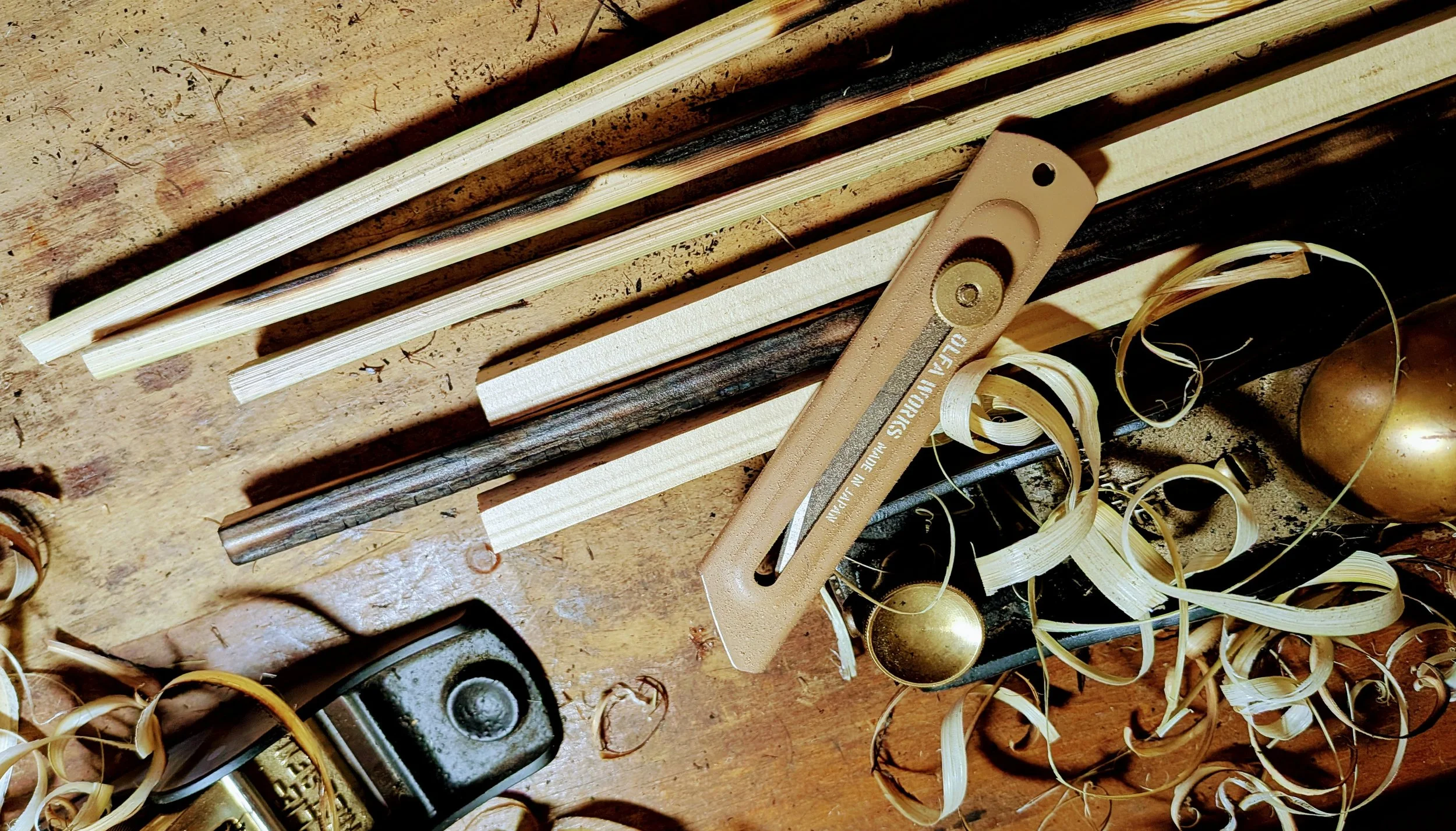Various wooden sticks, shavings, and shims on a workbench, with a wood chisel and part of a brass faucet in the background.