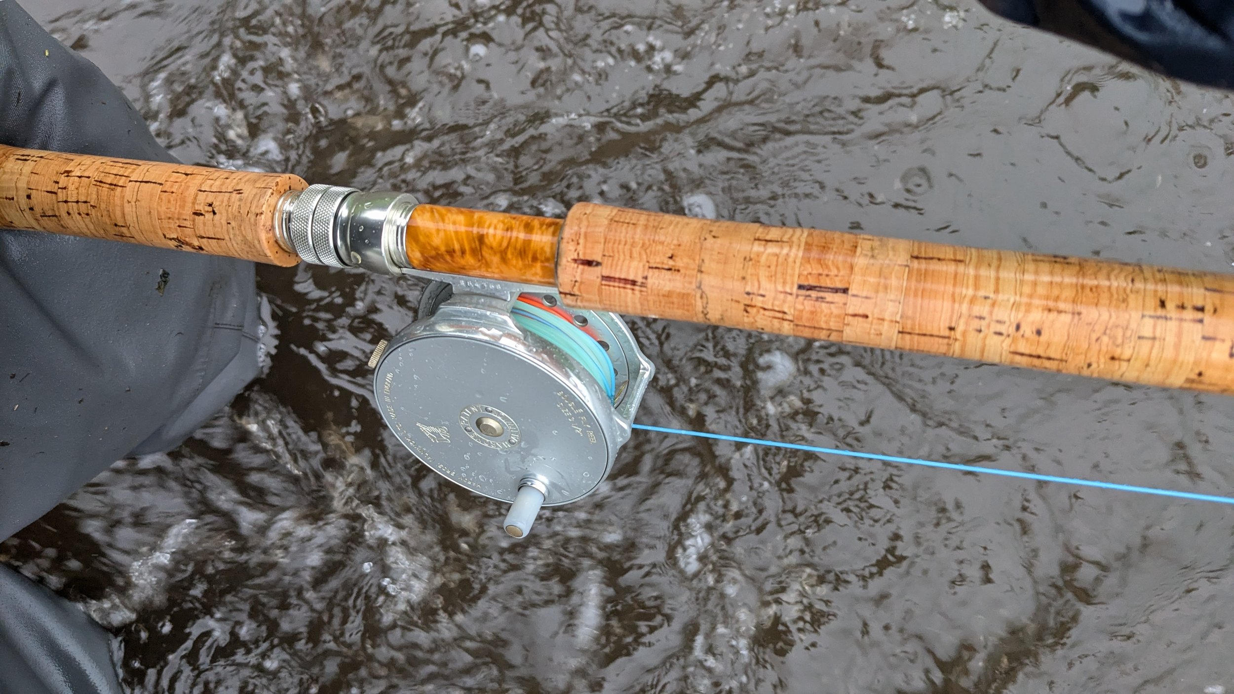 A fishing rod with a cork handle and spinning reel is held over a body of water with ripples and small waves.
