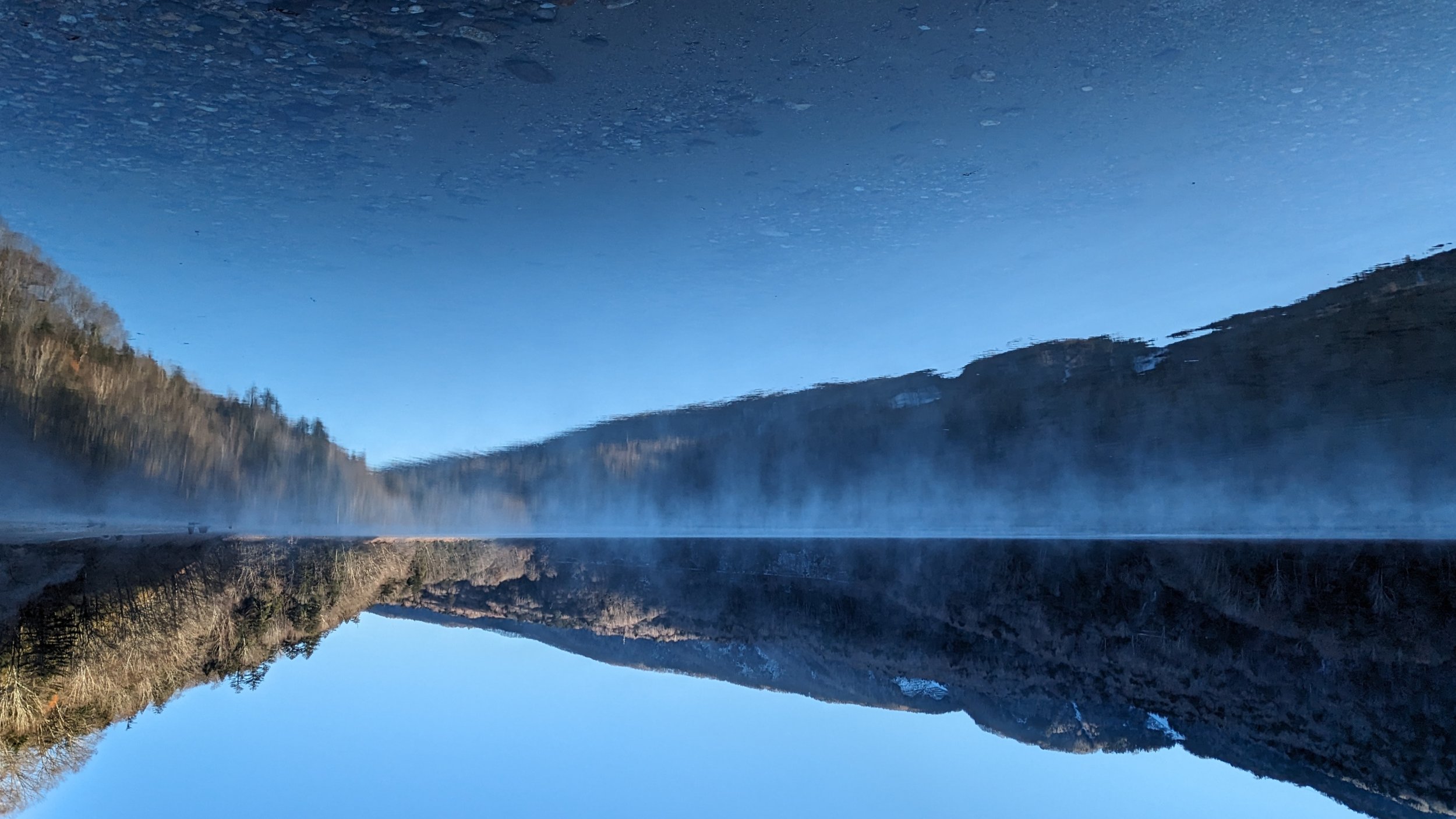 Reflection of mountains and clear blue sky on a calm lake with mist rising from the water.