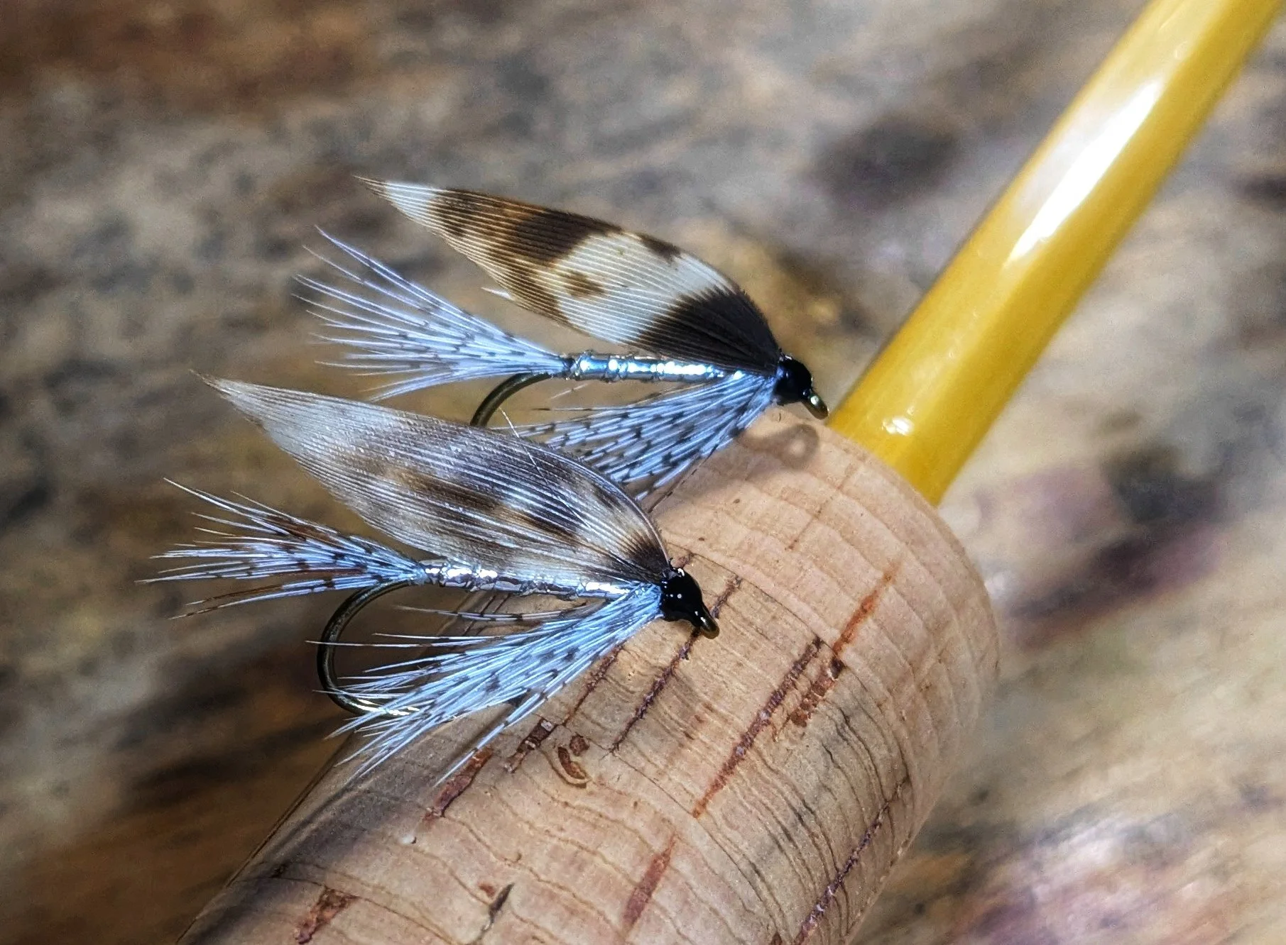 Close-up of three fishing flies with black and white patterned wings attached to a wooden handle, on a blurry brown background.