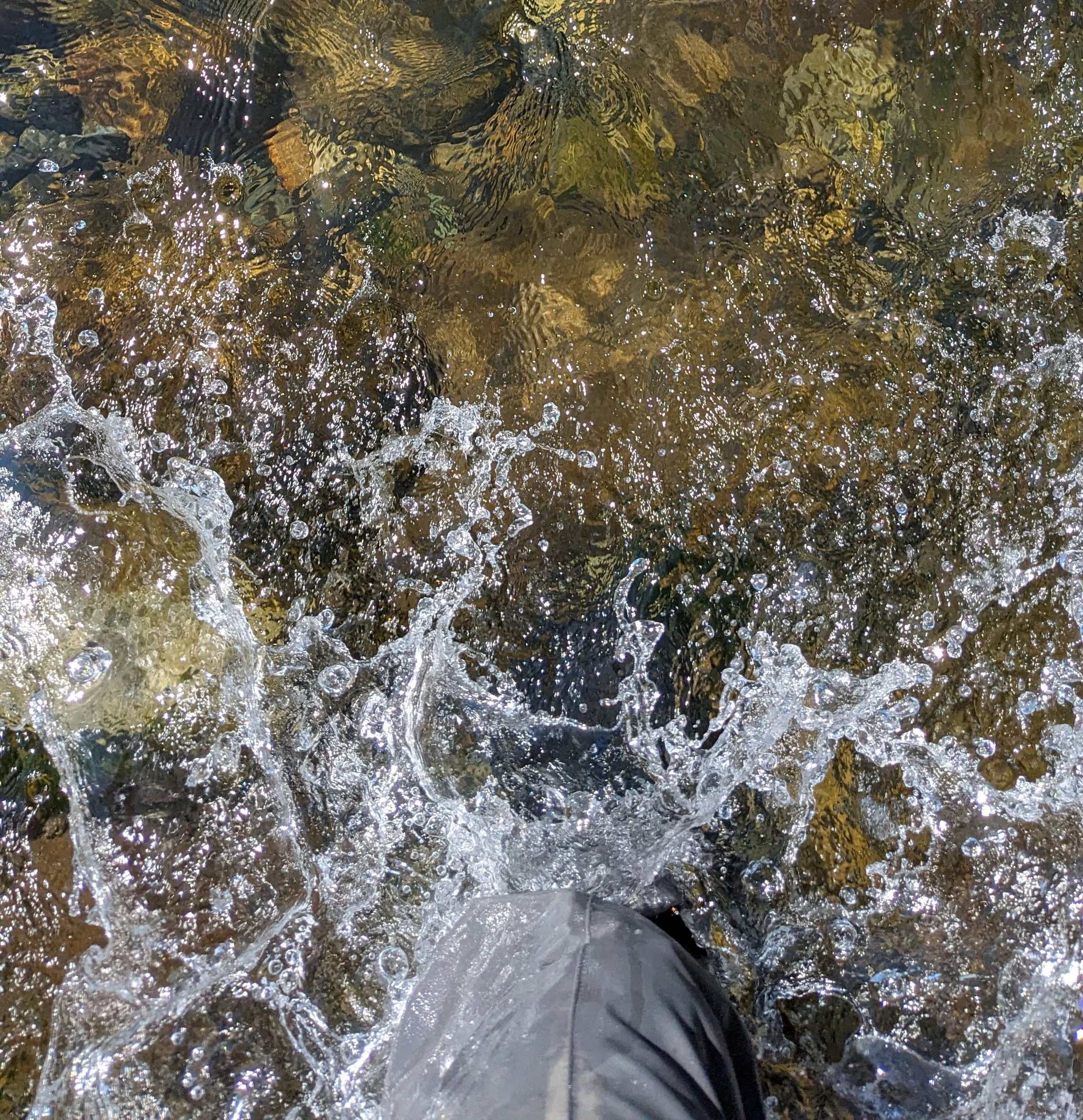 A person wearing black shoes standing on wet rocks near water, with sunlight reflecting off the water's surface.