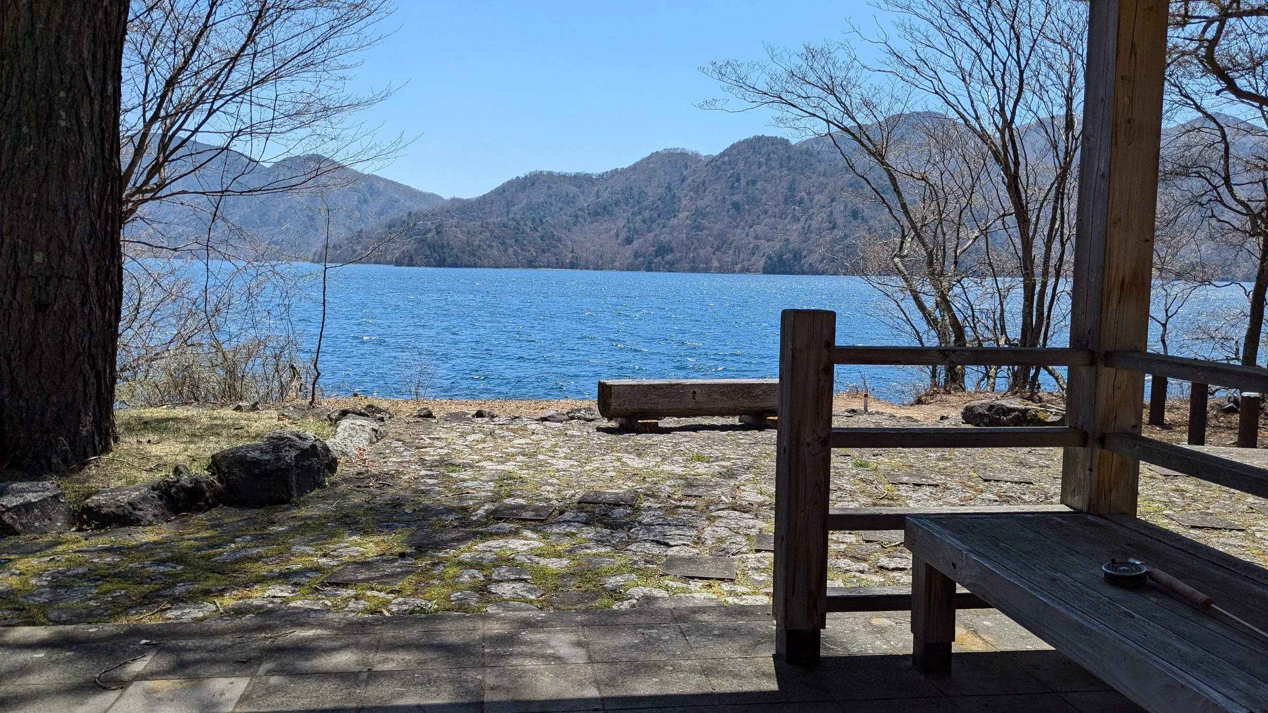 View of a large lake with mountains in the background, framed by leafless trees and a wooden deck with a bench in the foreground.