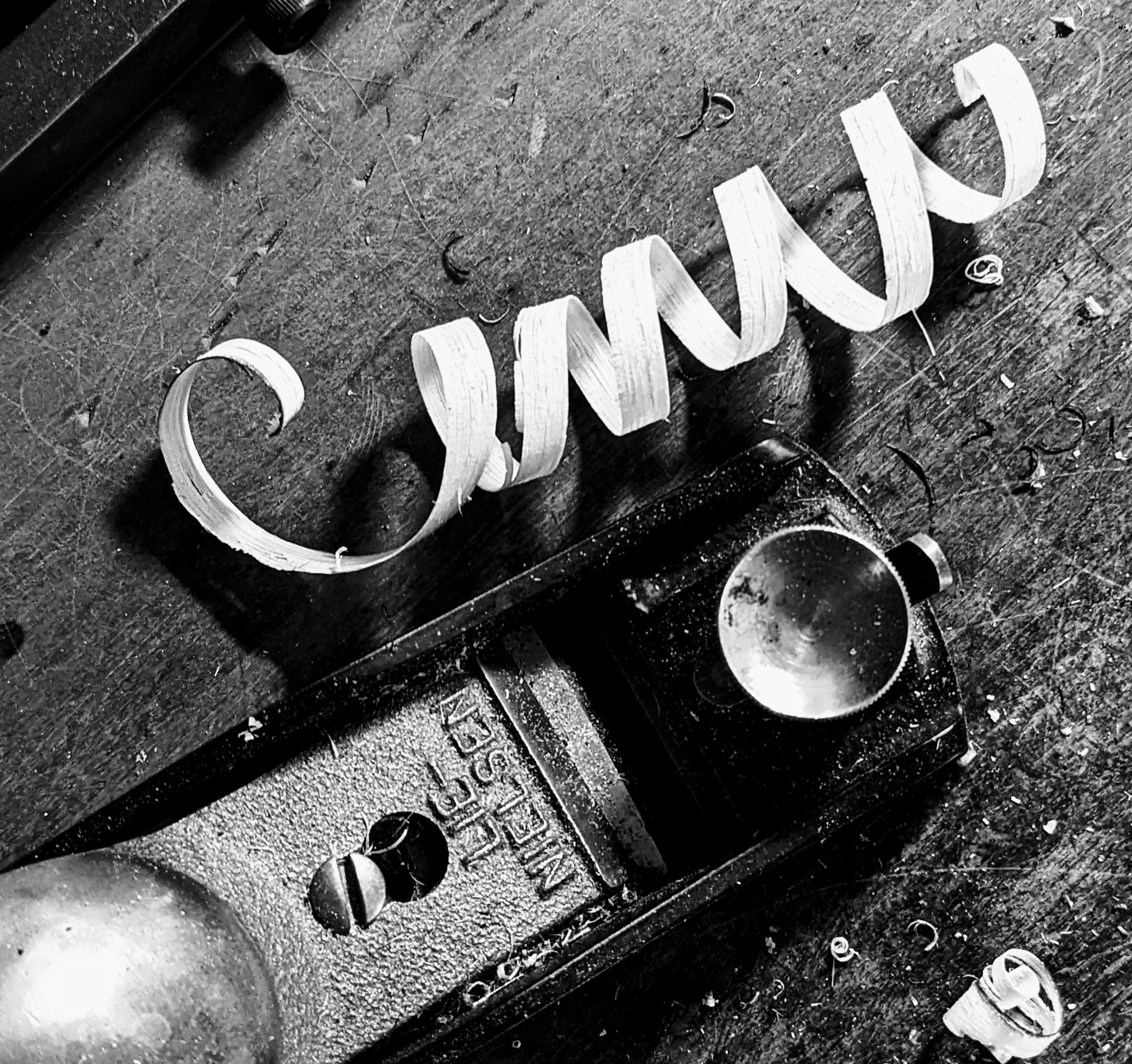 Close-up photo of a wooden sign lying on a rough, textured surface. The sign appears to spell out the word 'CINEMA' in cursive lettering. Surrounding the sign are some small debris and a cup, with part of a person's shoe visible at the bottom right c