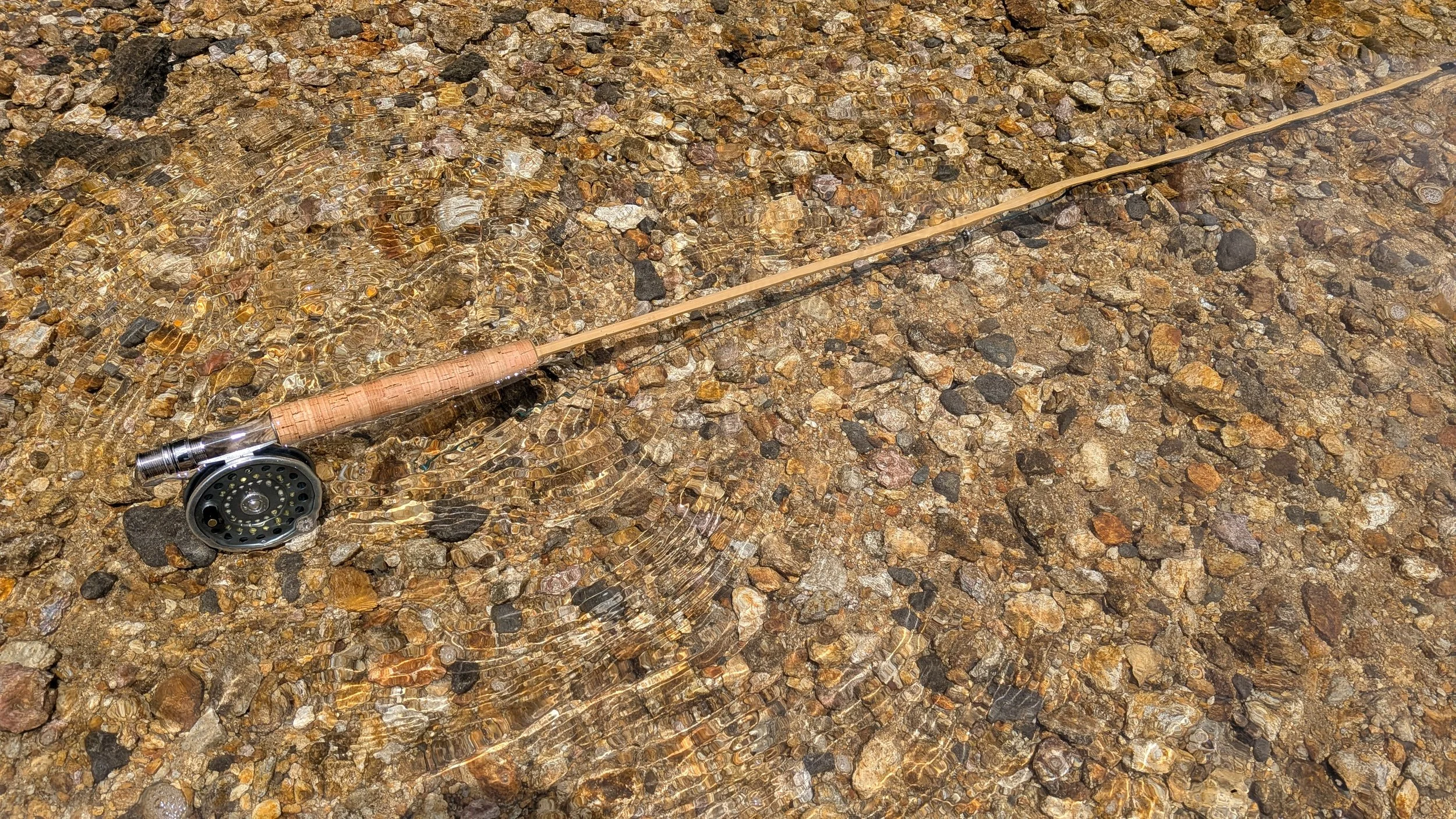 A fishing rod with a reel lying on a rocky shoreline partially submerged in clear water.