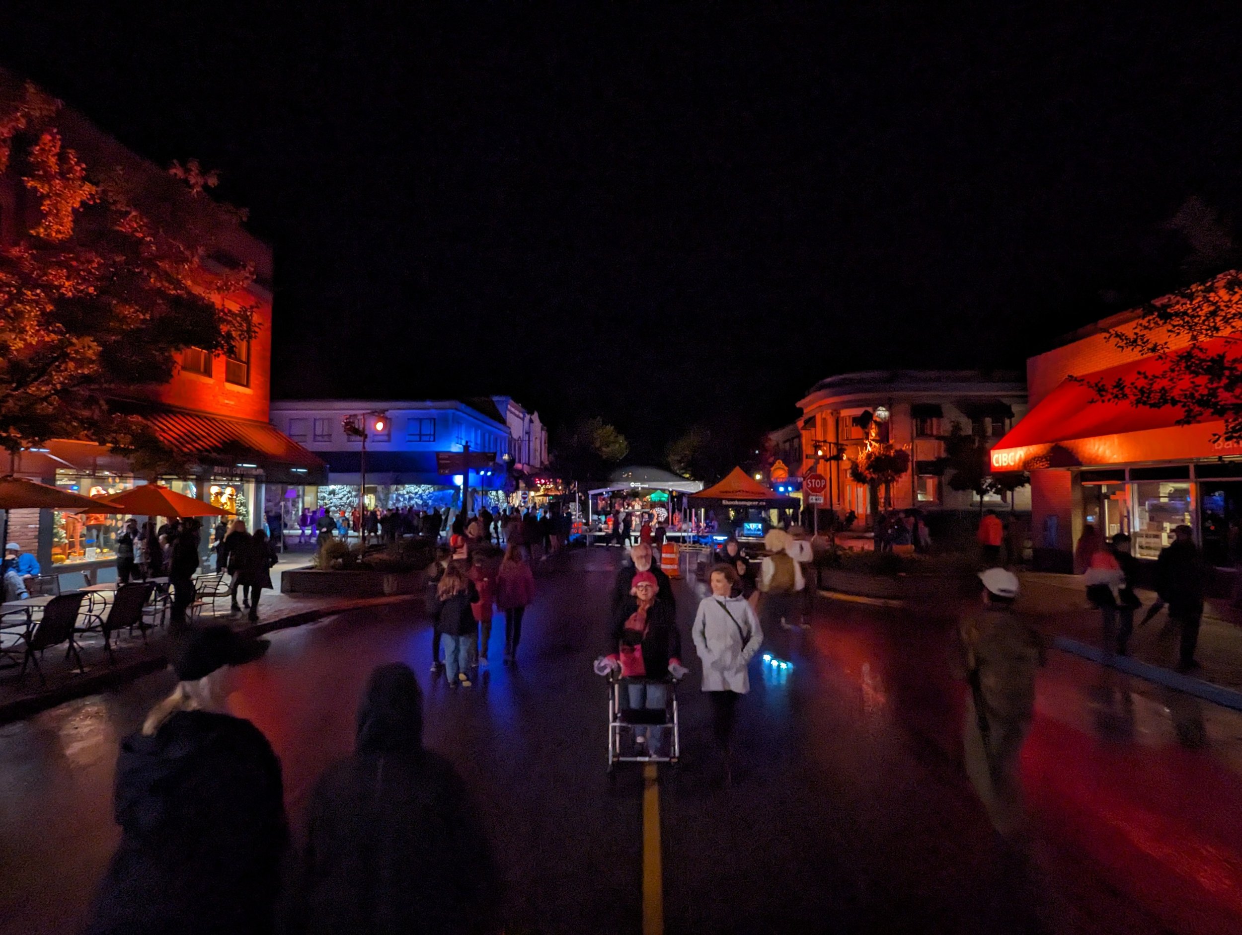 A nighttime street scene with people walking in a lively downtown area illuminated by colorful lights. The buildings are lit with red and blue lights, creating a festive atmosphere. There are shops and cafes with outdoor seating along the street.