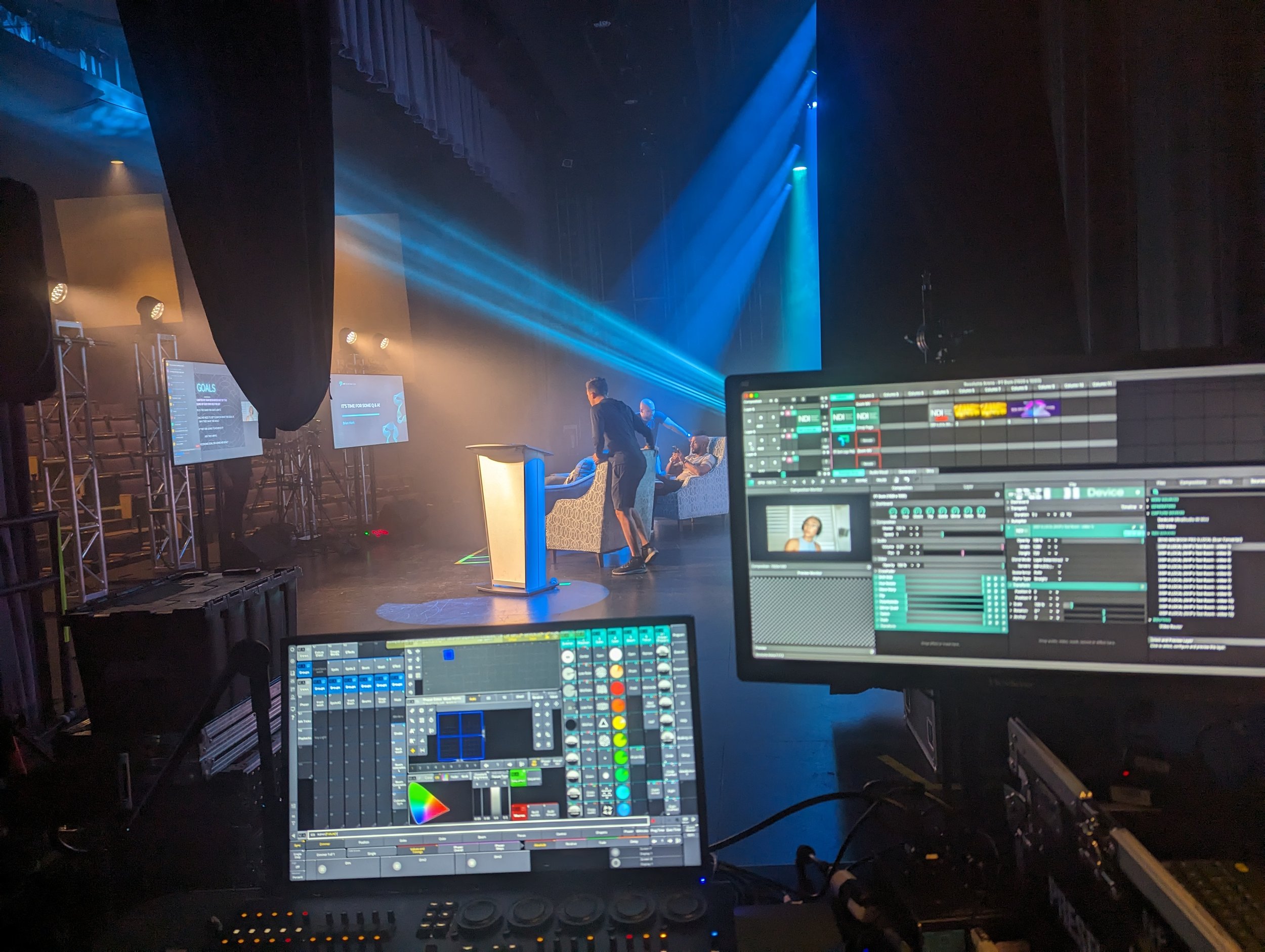 Backstage view of a stage setup with control monitors, lighting effects, and a presenter at a podium.