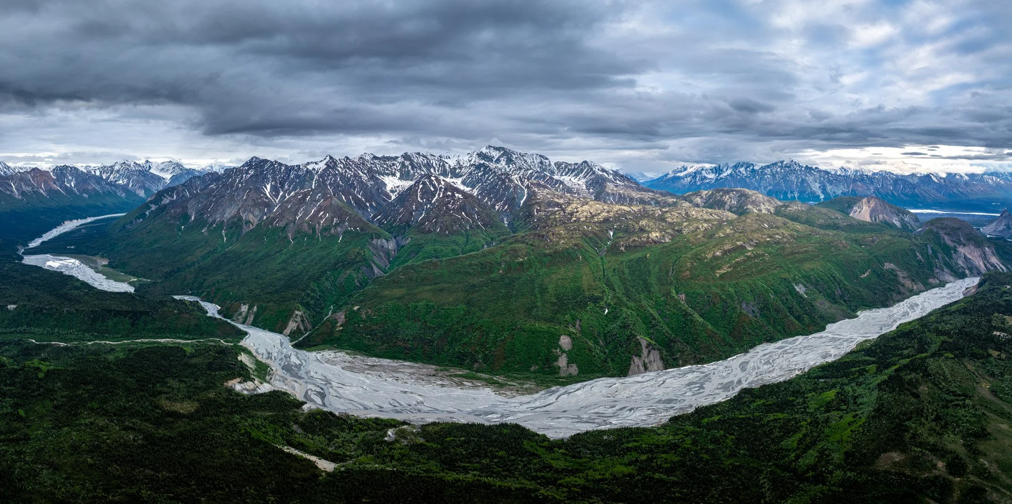 Mountain range with snow-capped peaks and green valleys, river flowing through the valley, overcast sky