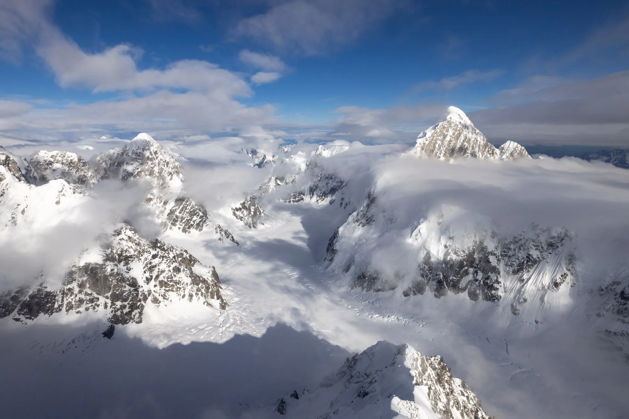 Snow-covered mountain peaks piercing through clouds under a blue sky.