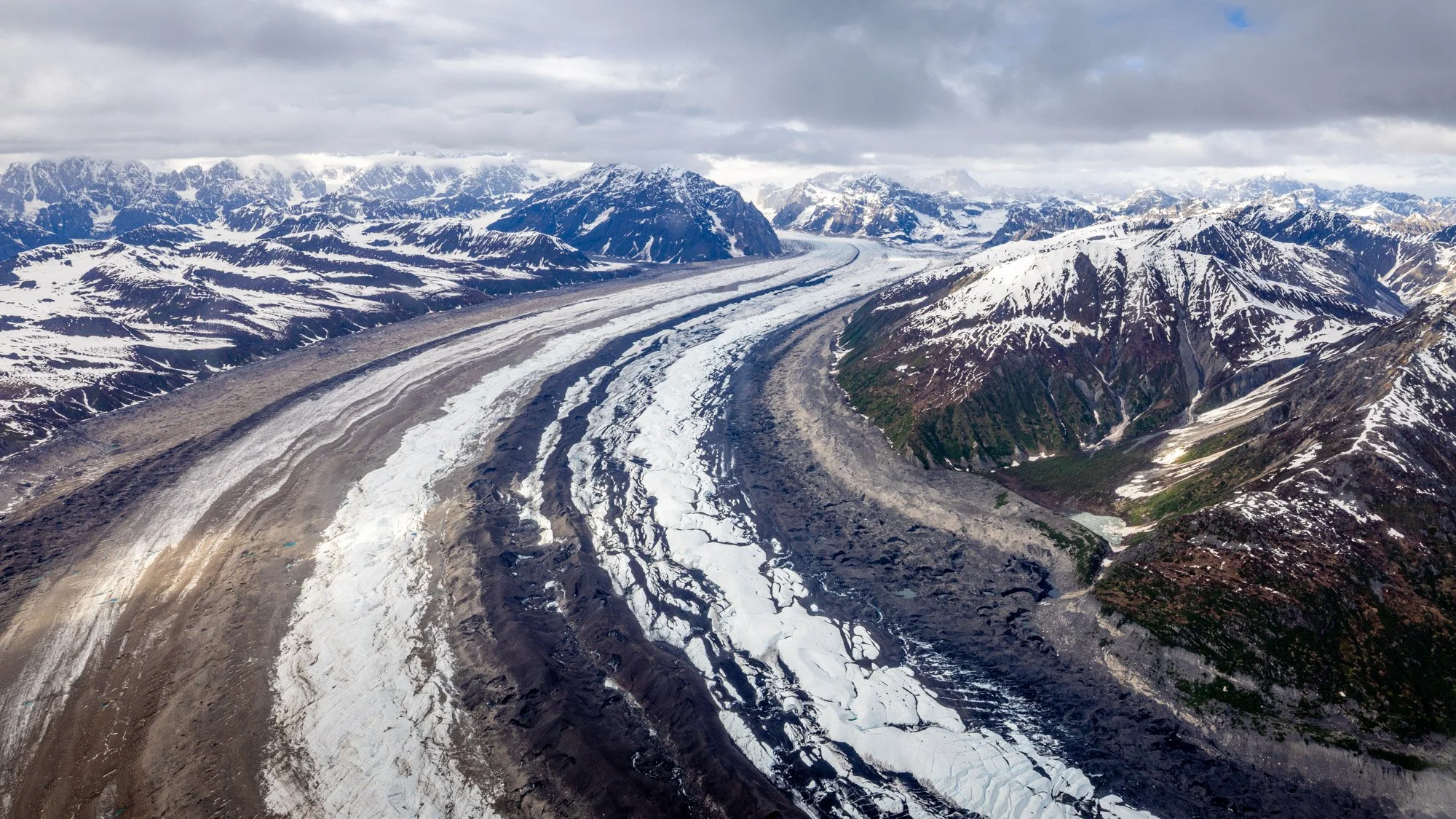 Aerial view of snow-capped mountains and glaciers with cloudy sky above.