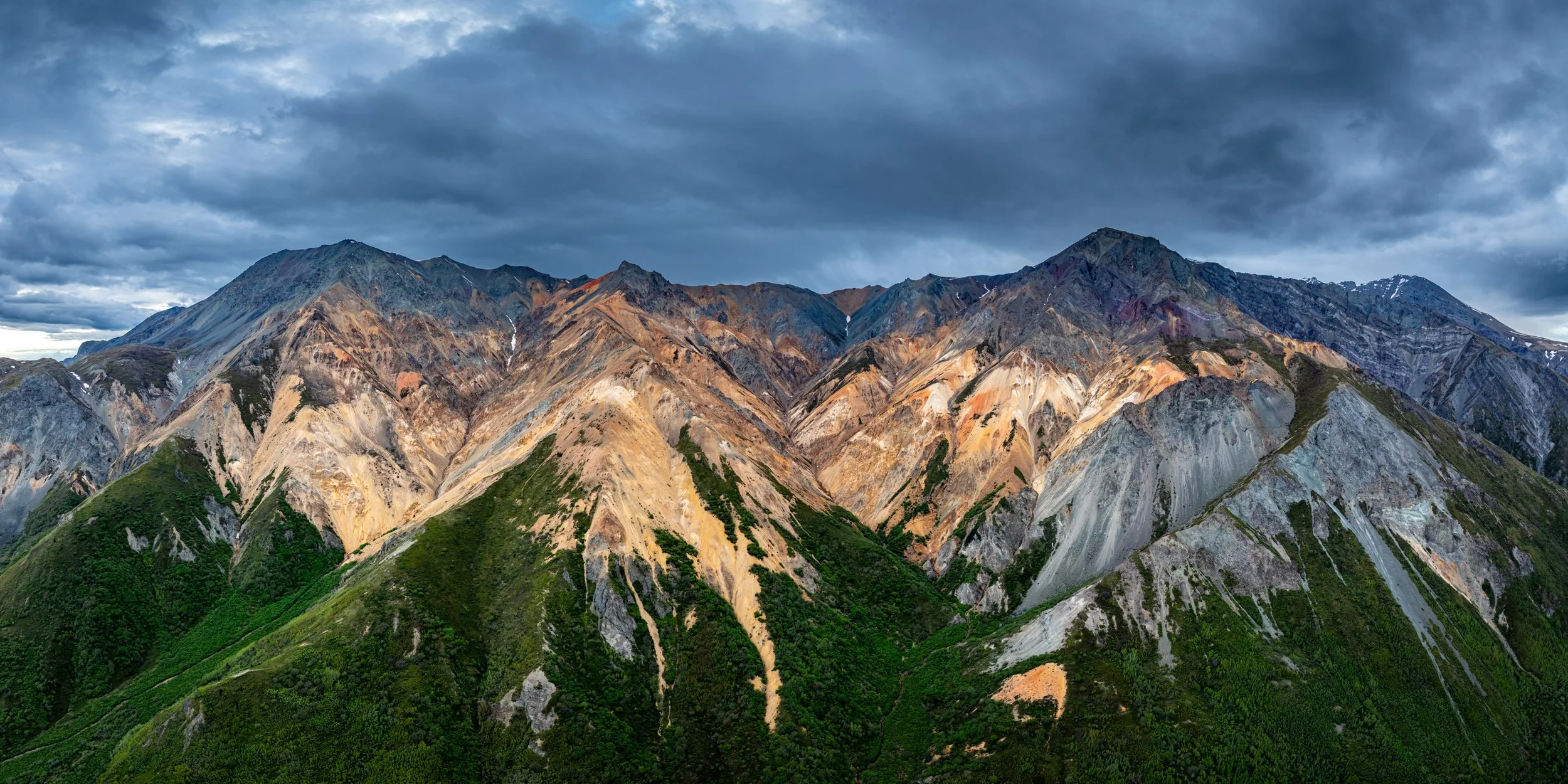A mountain range with colorful, jagged peaks under a dark, cloudy sky, with green vegetation at the base.