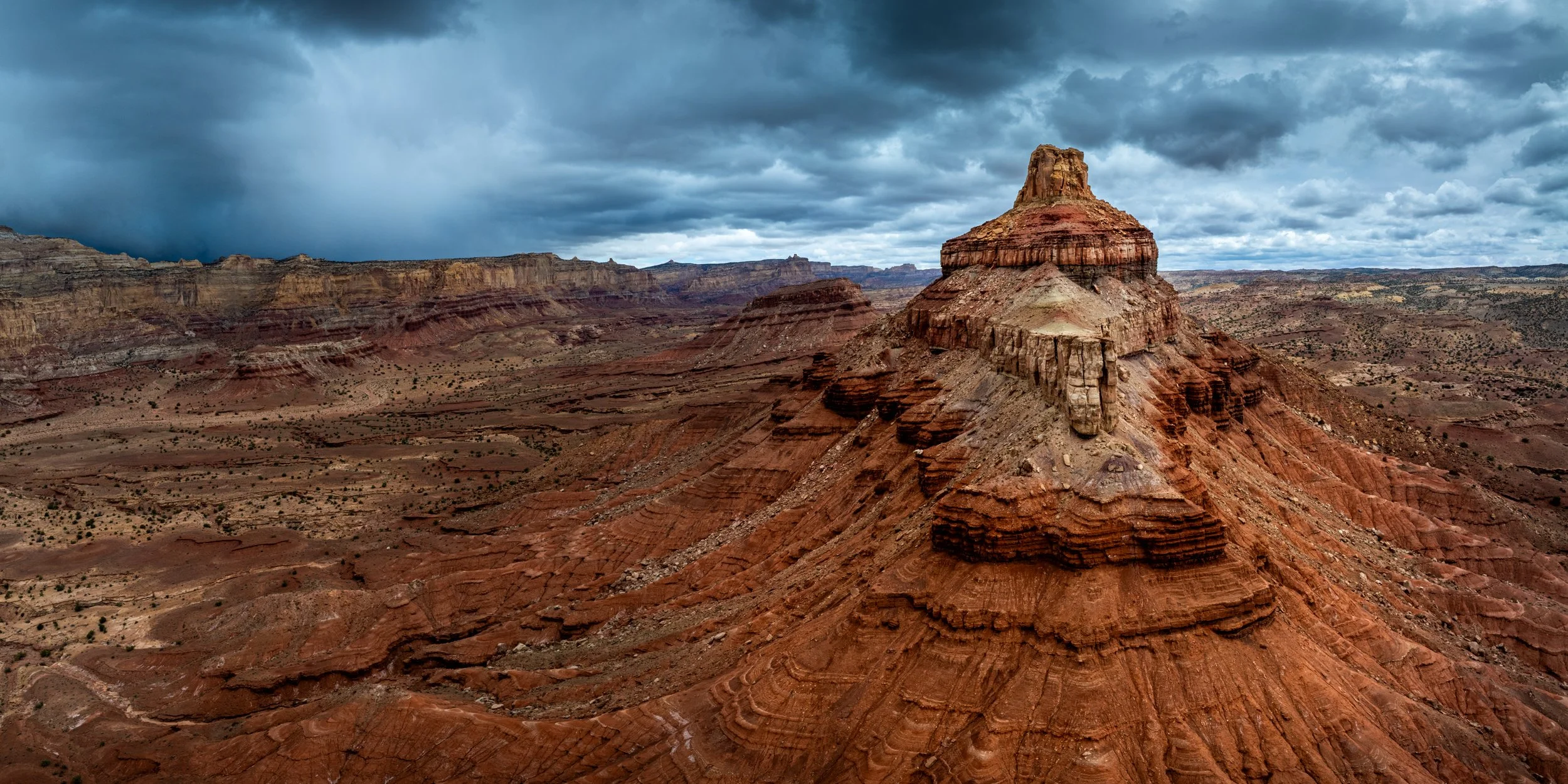 A rugged, reddish-brown rock formation in a desert landscape with a layered, flat-topped peak under a cloudy, stormy sky.