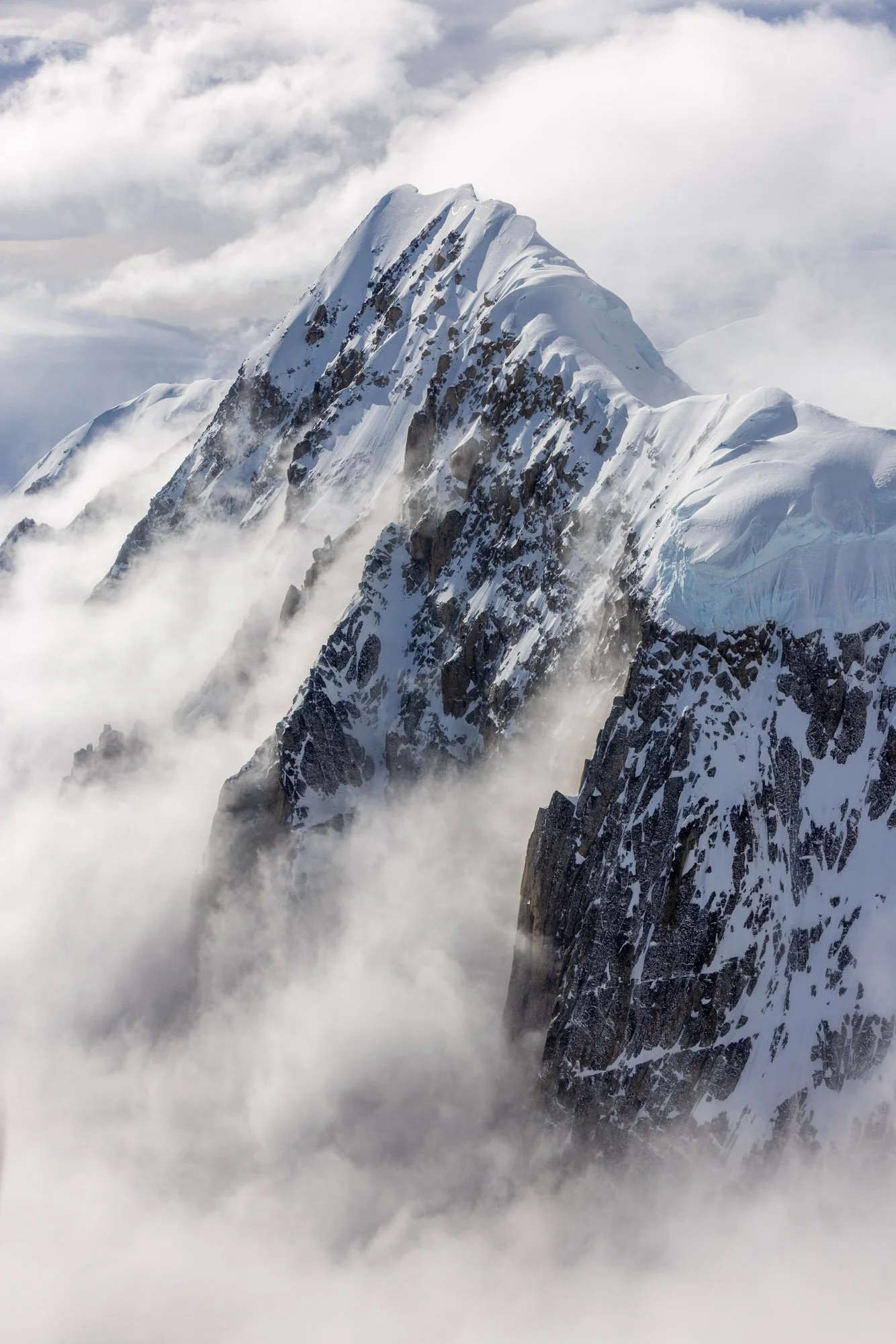Snow-covered mountain peak with rocky cliffs, surrounded by clouds and mist.