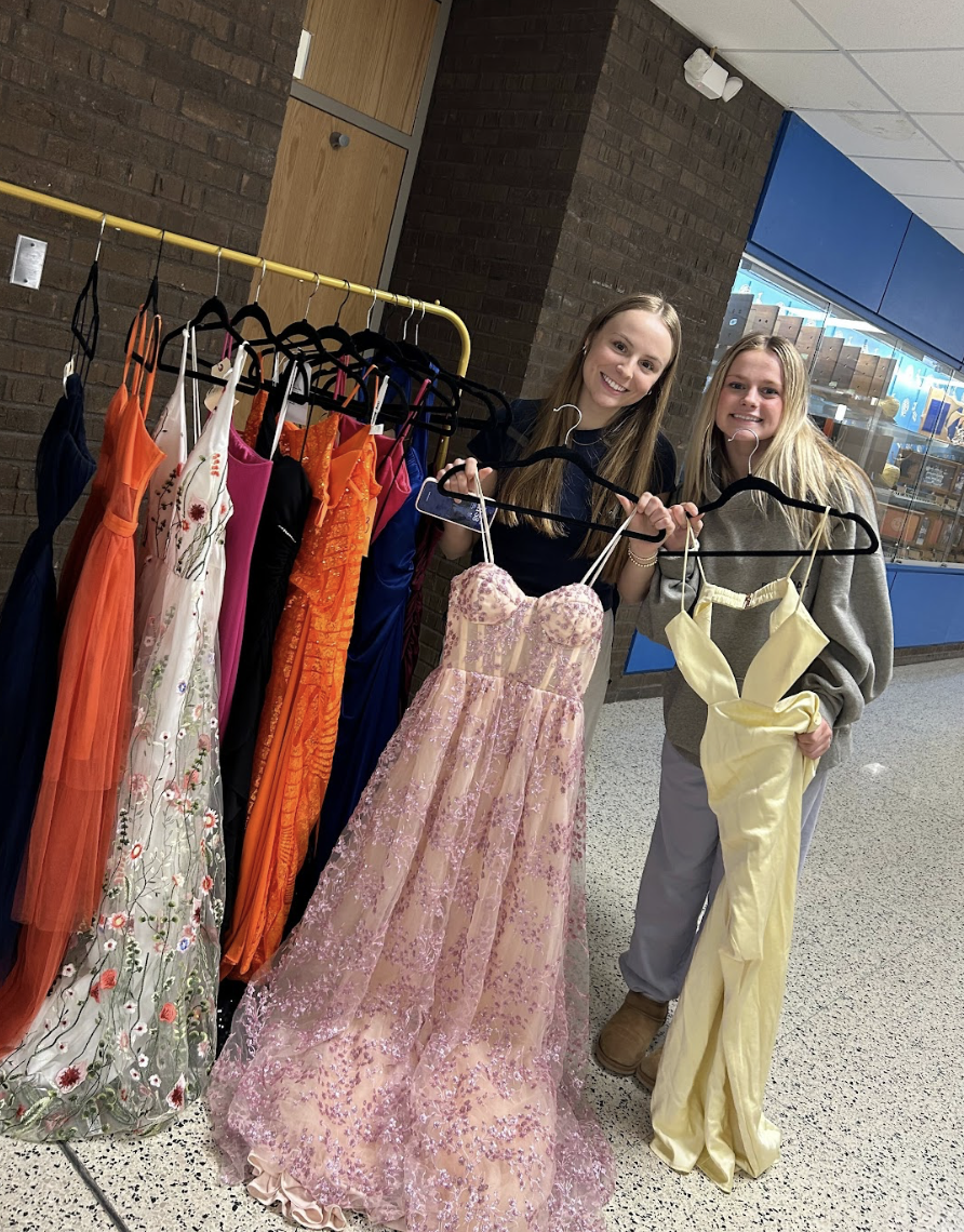 Two young women standing behind a clothing rack filled with colorful dresses, holding and smiling at elegant dresses, in an indoor space with brick and blue walls.