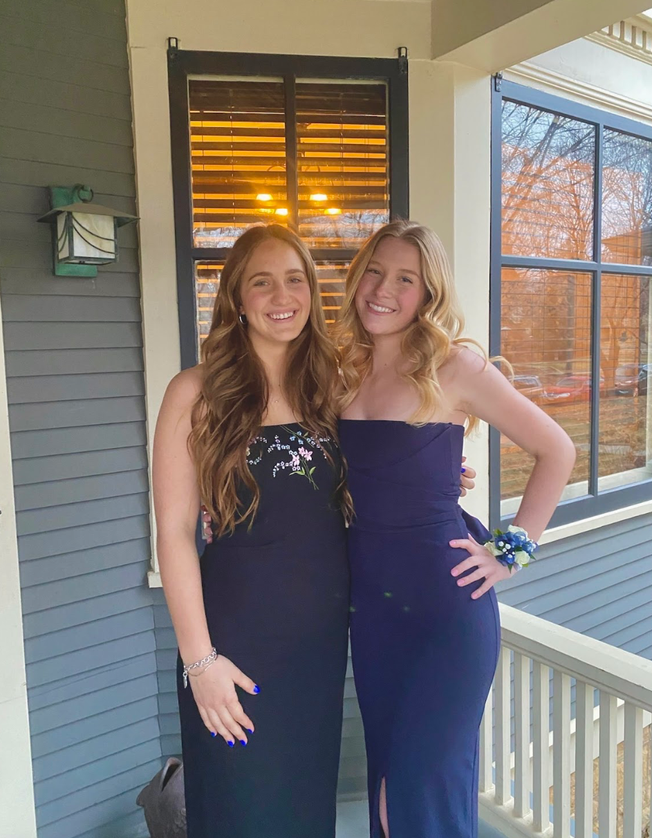 Two young women in navy blue evening dresses standing on a porch, smiling at the camera, with one wearing a corsage and the other a bracelet.