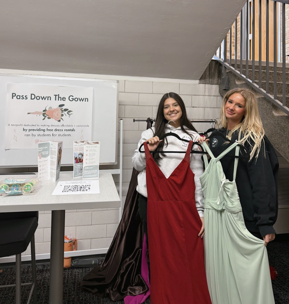 Two young women holding dresses in front of a sign that reads "Pass Down The Gown" promoting free dress rentals for students, with tables and informational materials nearby.