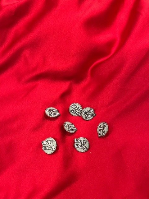 Small silver pins with the text 'Prom Mom' on a red fabric surface.