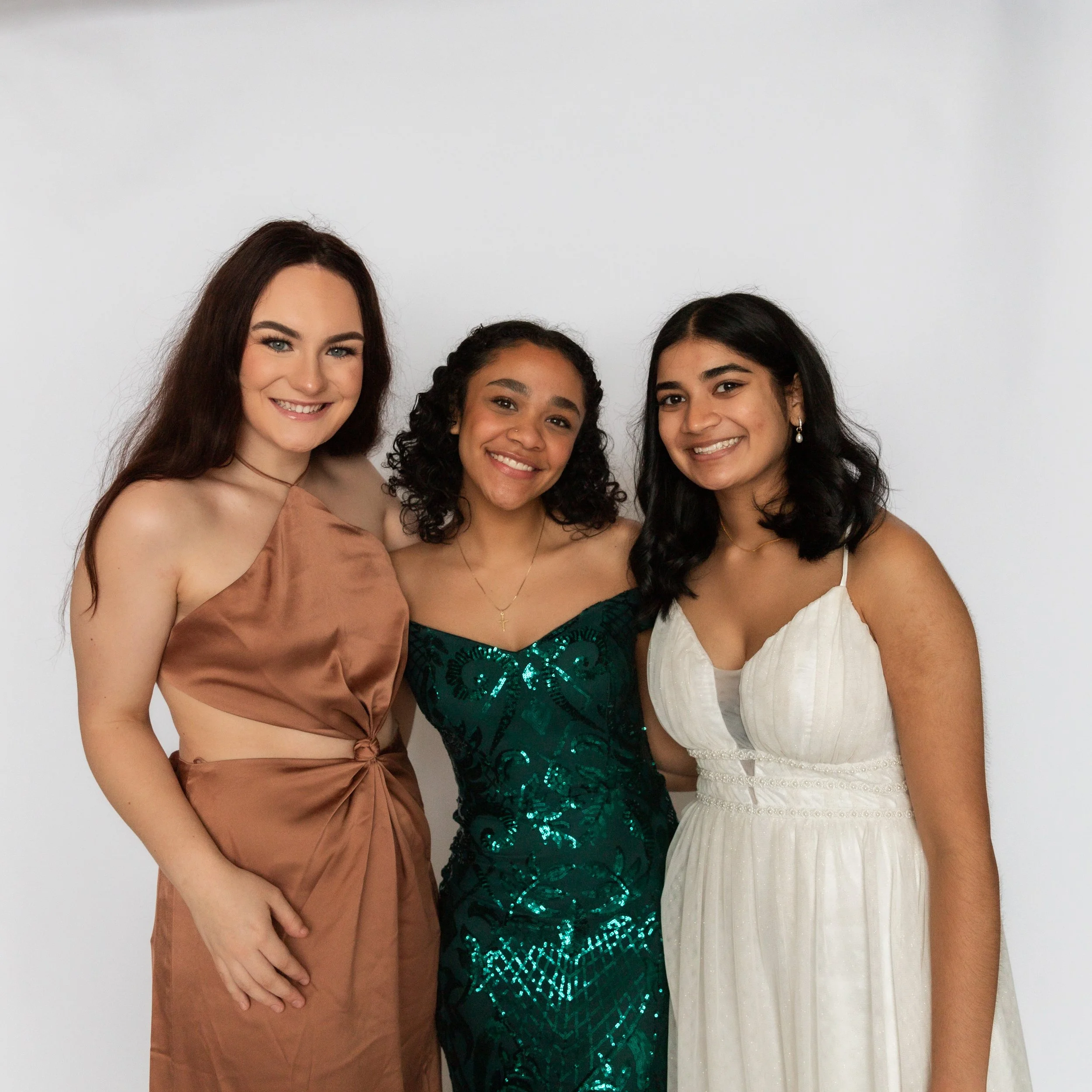 Three women smiling and standing close together in dresses against a plain white background.