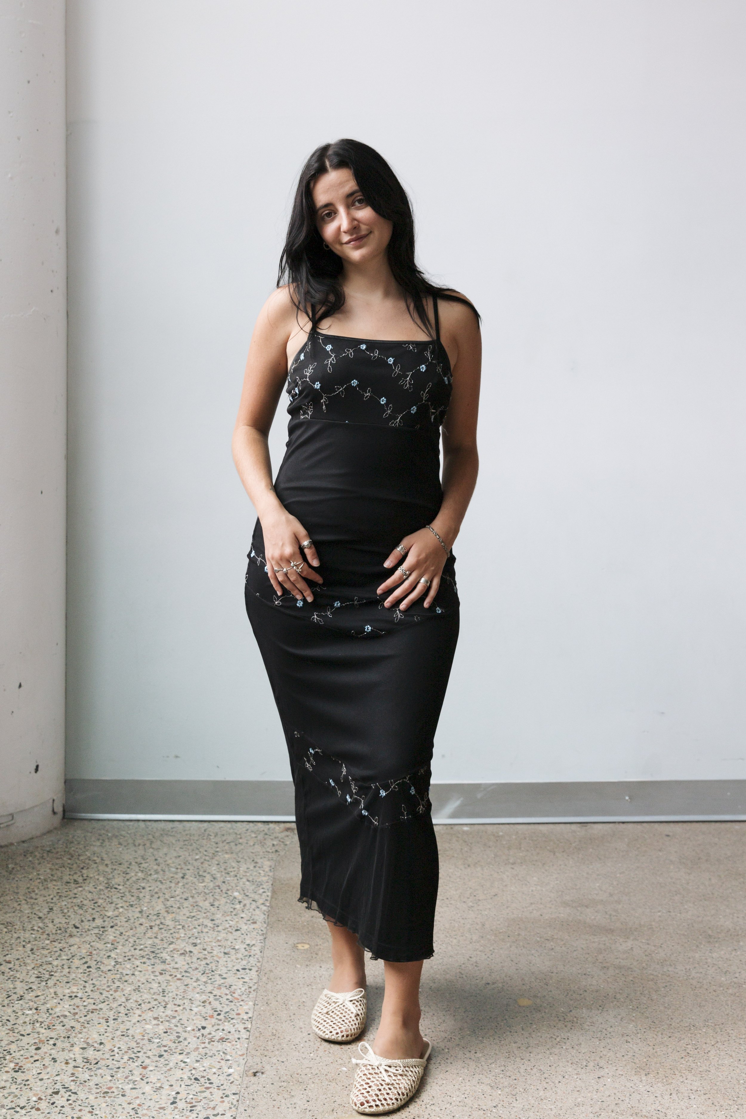 A young woman with dark hair wearing a black spaghetti strap dress with embroidered floral details and beige woven slip-on shoes standing against a plain white wall.