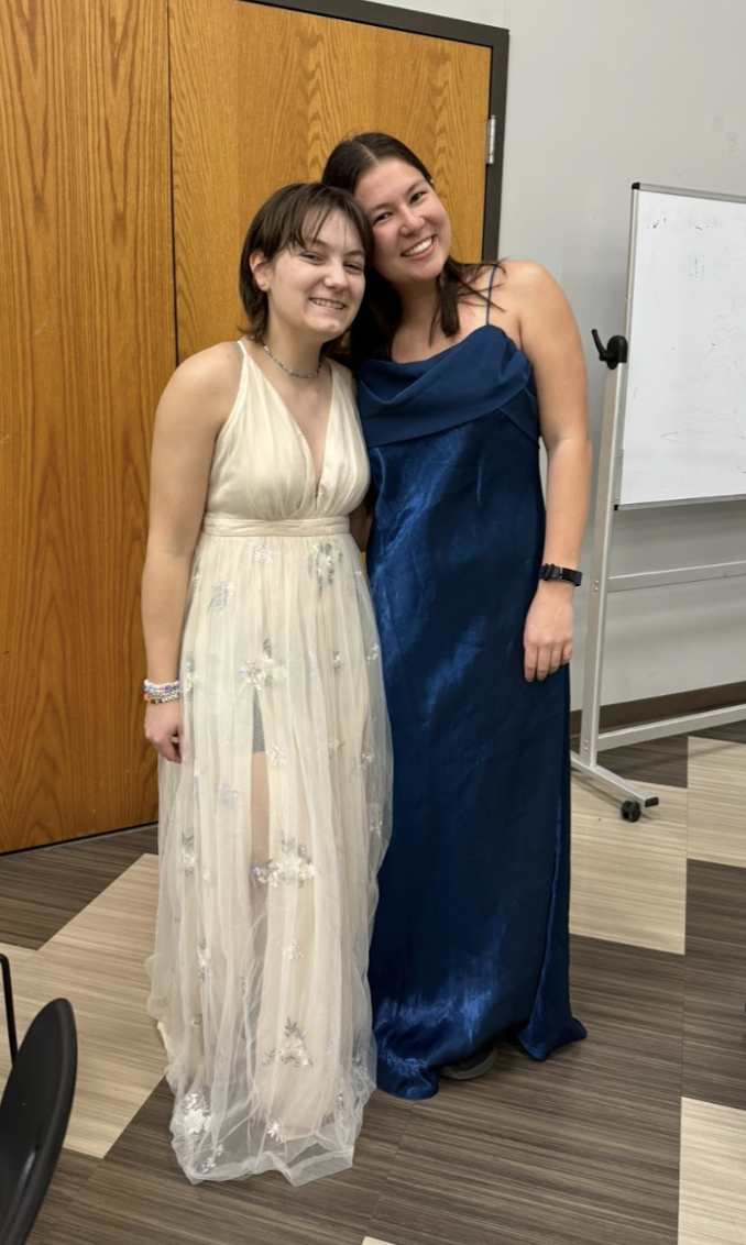Two women standing close together, smiling, in a room with wooden doors and a whiteboard. One is wearing a cream-colored dress with floral details, and the other is wearing a dark blue dress.