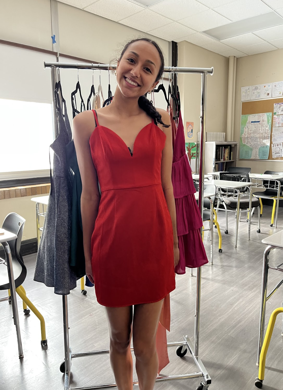 Young woman in a red dress standing in a classroom with dresses on a rack behind her.