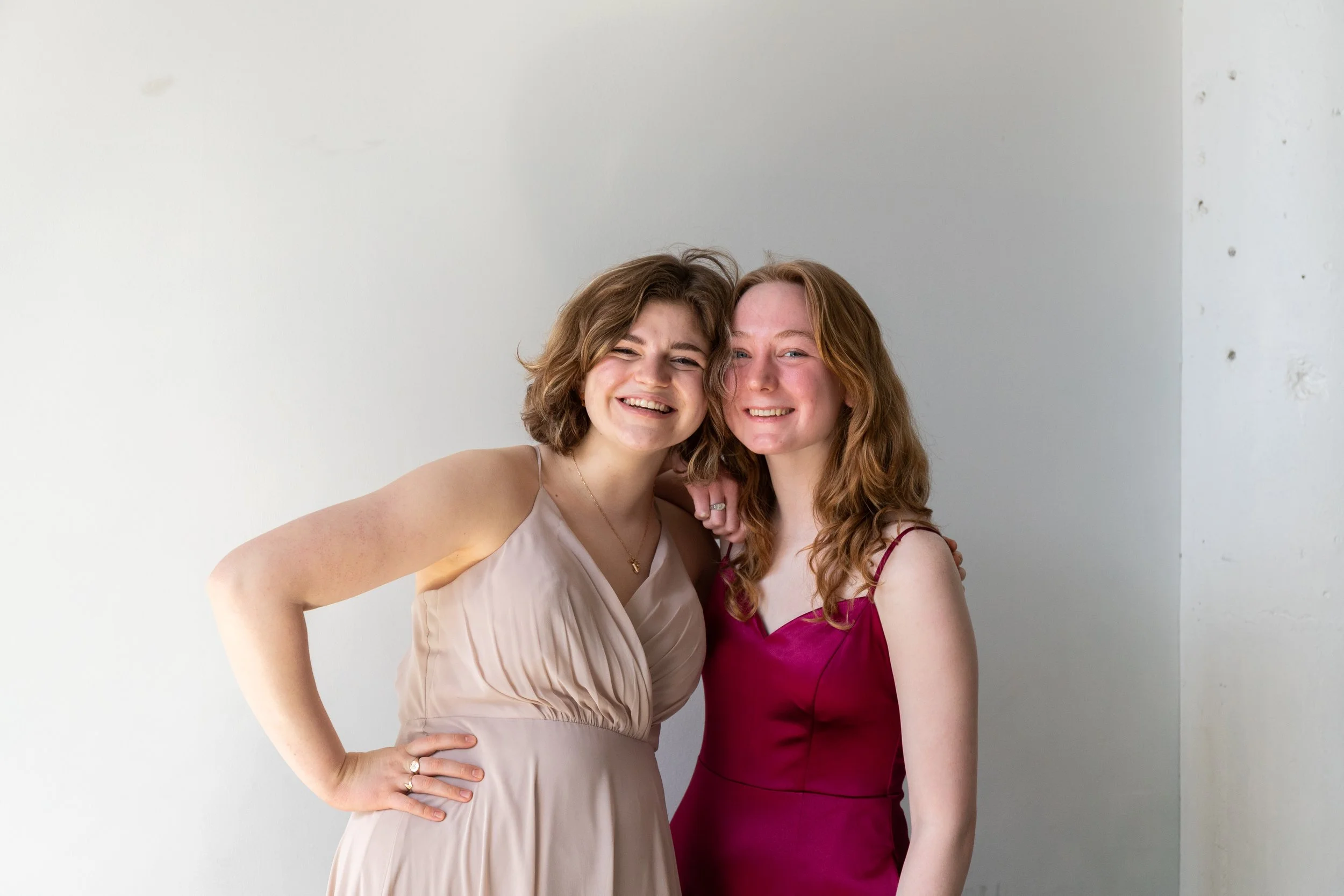 Two women with red and brown hair smiling and posing together, wearing elegant dresses, against a plain white wall.