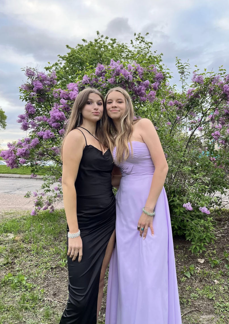 Two young women in elegant dresses standing outdoors in front of a flowering bush with purple blossoms, smiling at the camera.