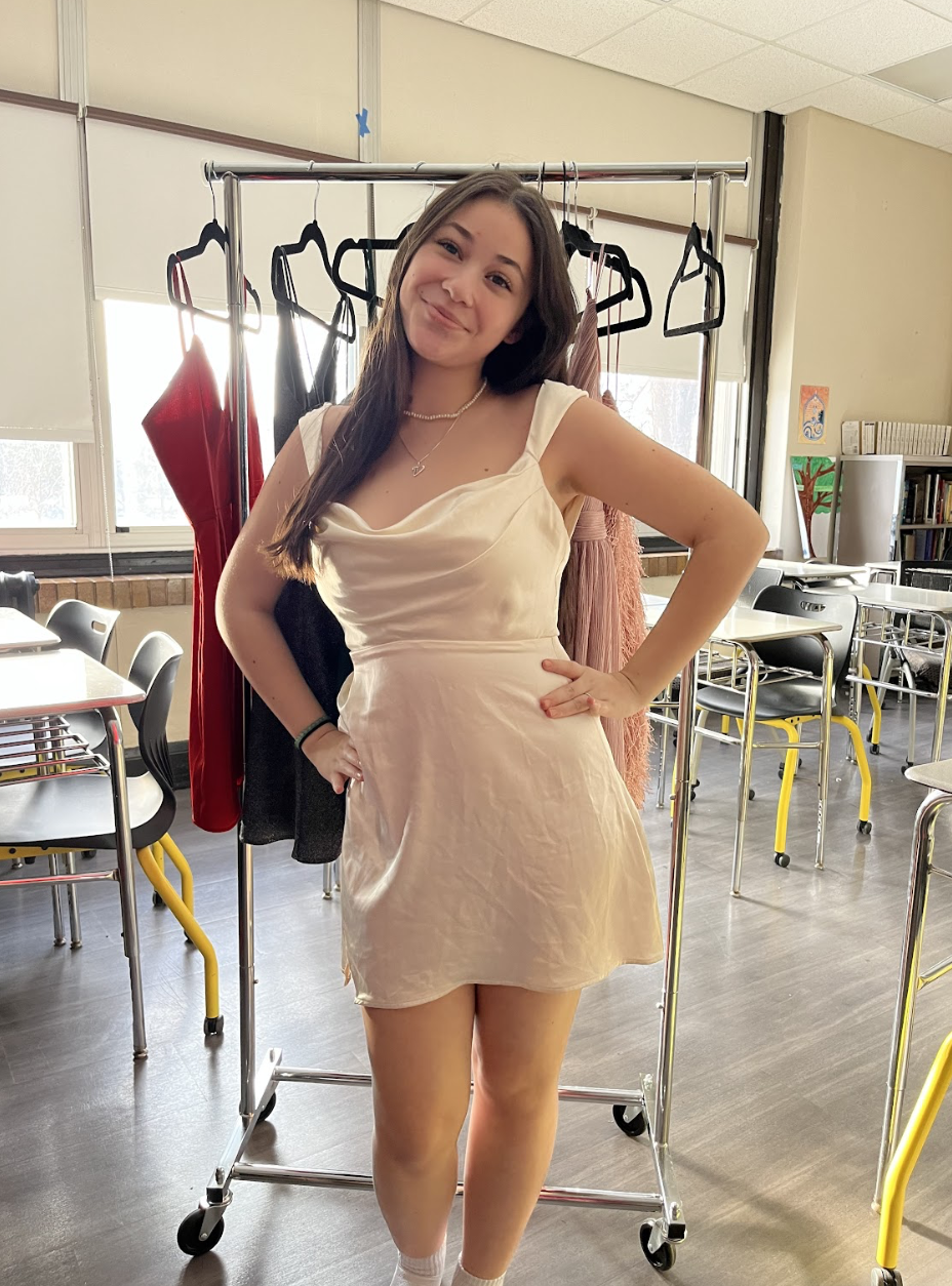 A young woman in a cream-colored satin dress standing in a classroom, smiling with her hands on her hips, with a clothing rack behind her holding various dresses.
