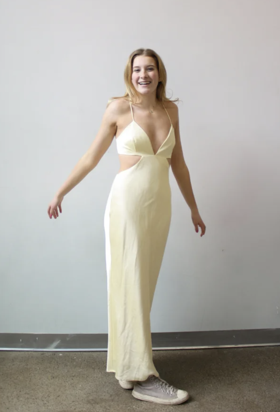A young woman in a yellow cut-out dress and white sneakers standing against a plain light gray wall, smiling.